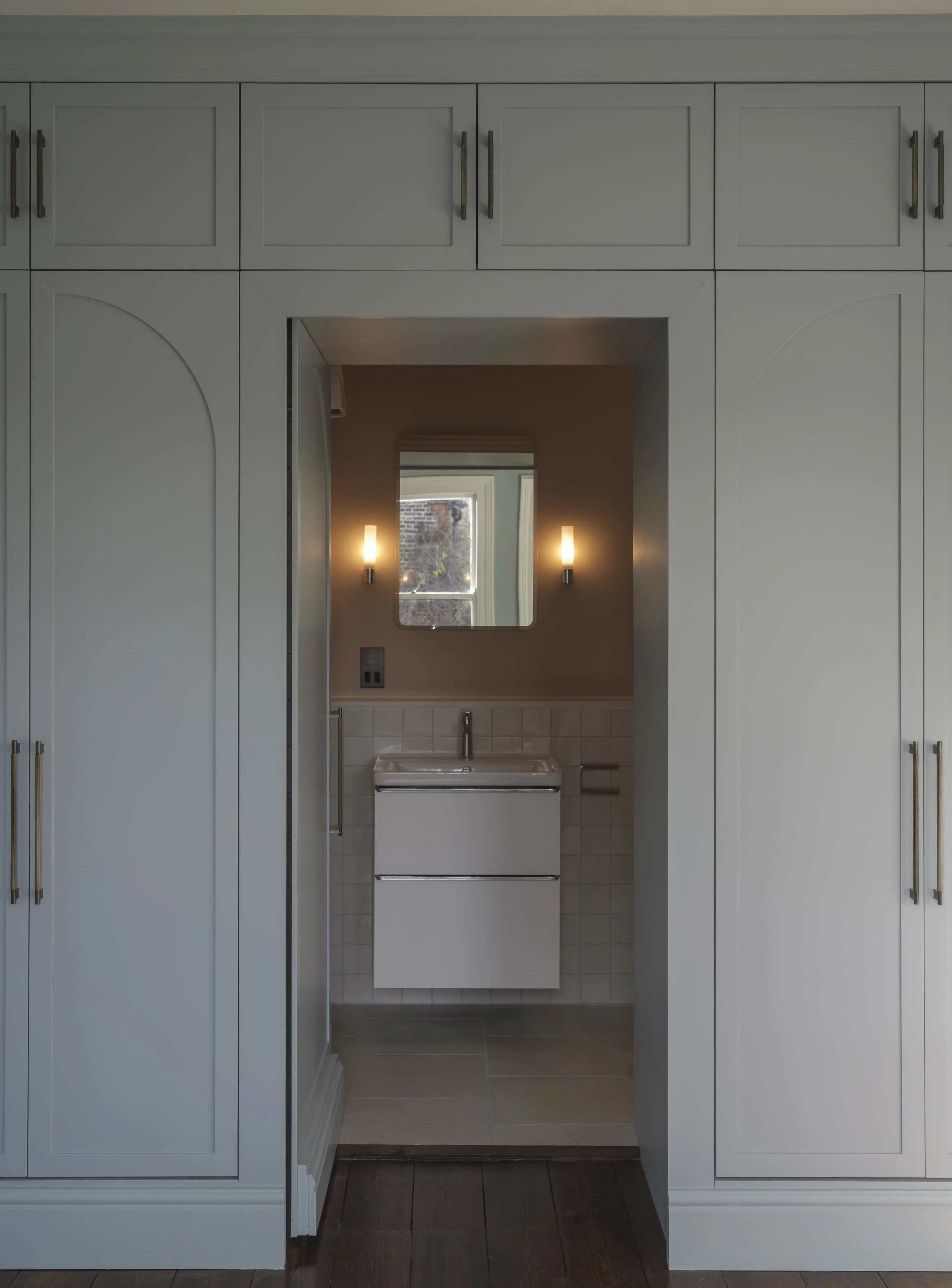 A kitchen with light blue cabinets and a small bathroom area visible through a doorway, featuring a mirror, two wall-mounted lights, and a window showing an outside brick wall.