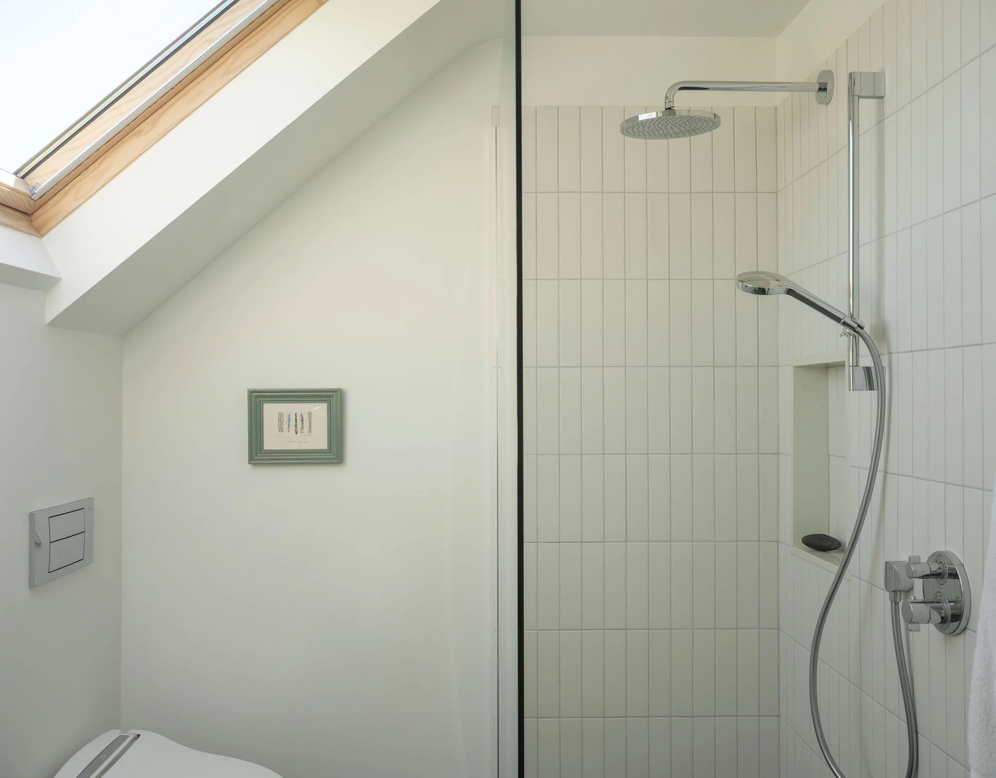 A modern walk-in shower with a large rain showerhead, a handheld showerhead, white tile walls, and a black trim in a bright bathroom with a sloped roof and a skylight window.