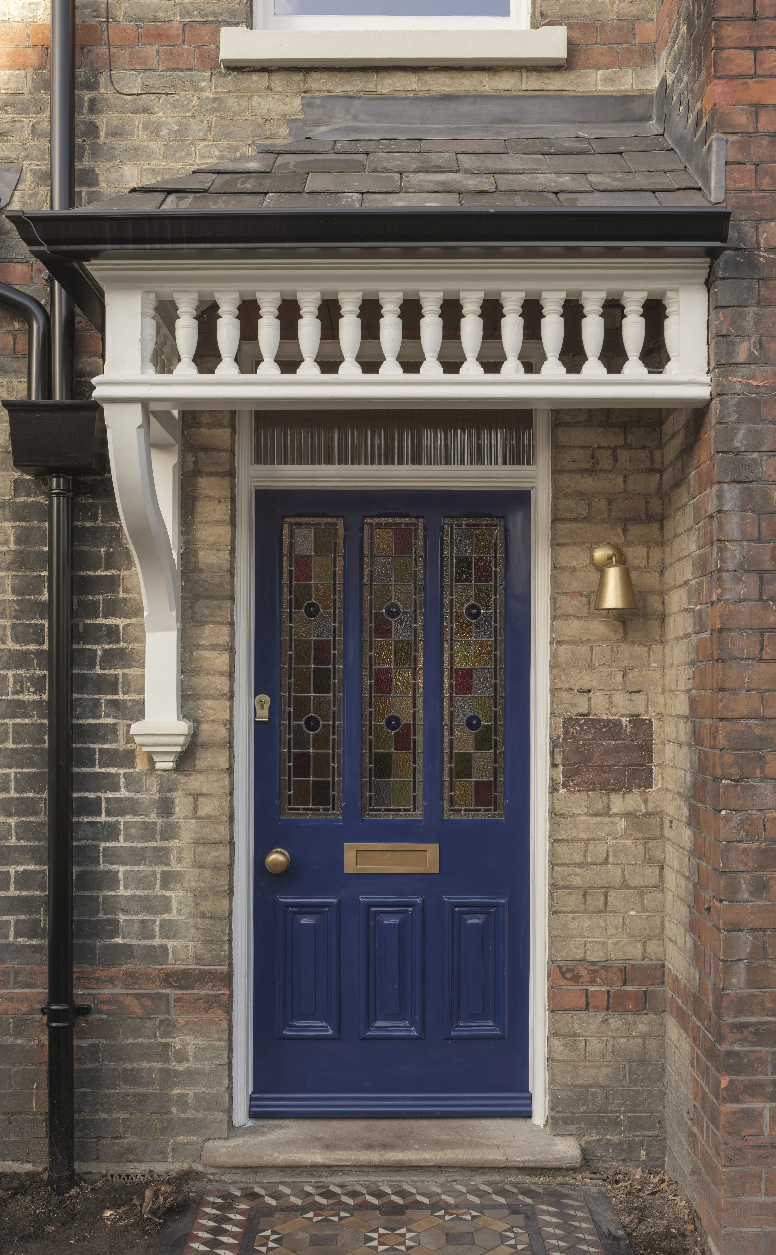 Blue front door with stained glass panels, brass mail slot, and doorknob, framed by brick walls and a tiled porch area, with a small brass wall-mounted light fixture to the right of the door.