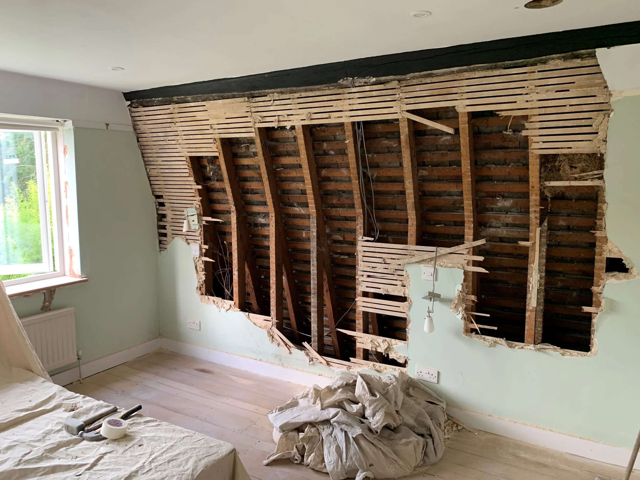 Room under renovation with plaster removed from wall, exposing wooden studs and brickwork, with plastic sheets and tools on the floor.