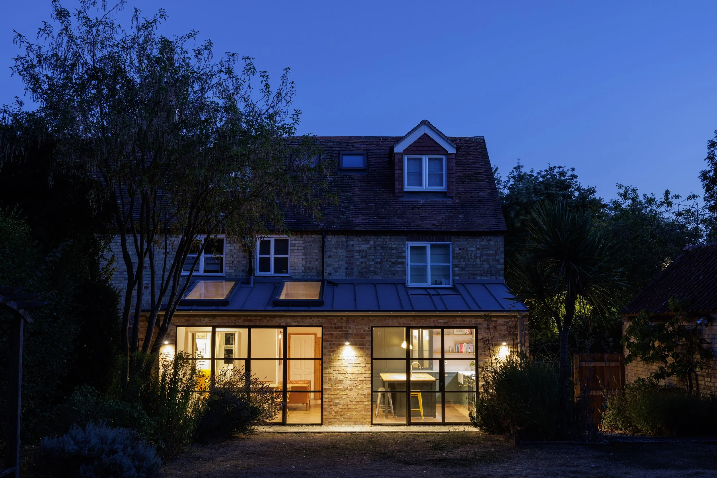 The image shows a brick house at dusk with large windows illuminating the interior, surrounded by trees and plants.