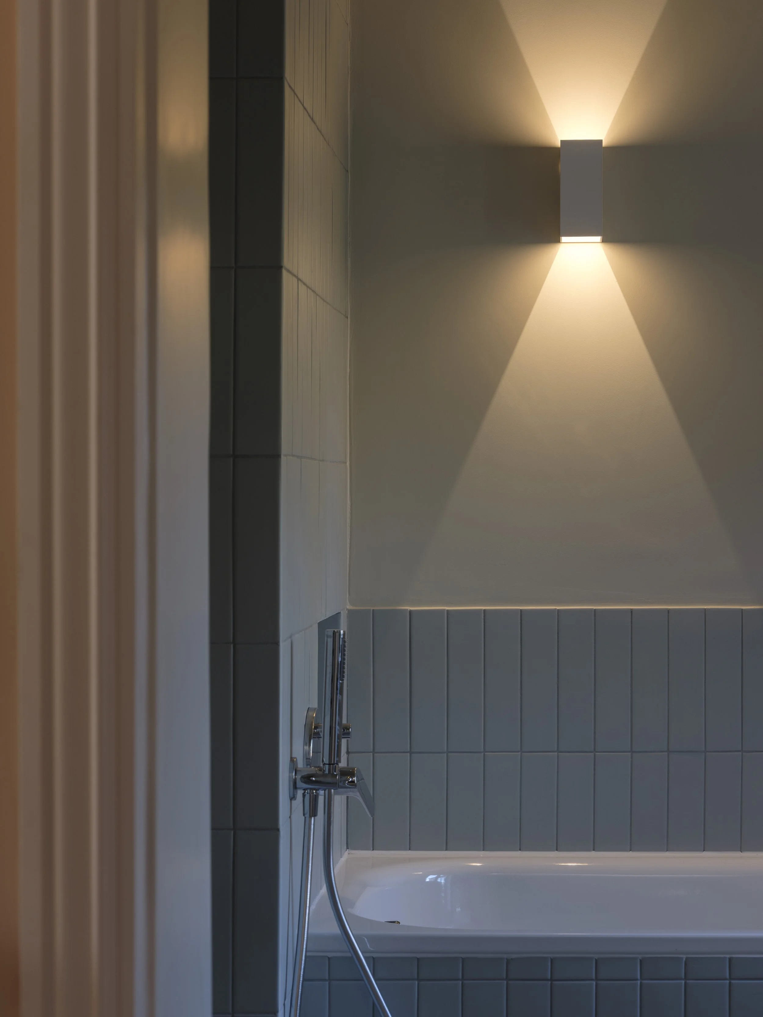 Interior of a bathroom with a bathtub, tiled wall, and a modern wall-mounted light fixture casting light upward and downward.