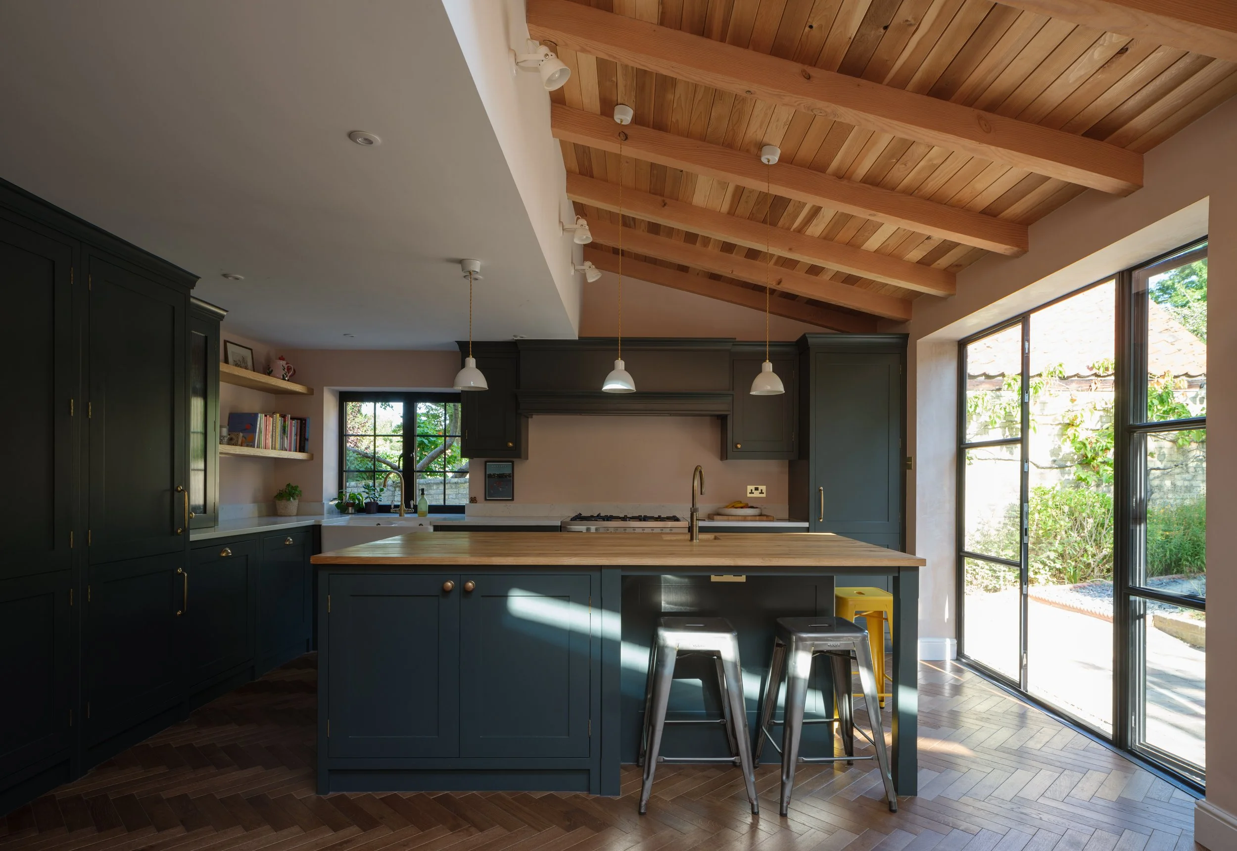 Kitchen with dark green cabinets, a wooden island, hanging pendant lights, and large windows with a view of greenery outside, featuring a sloped wooden ceiling.