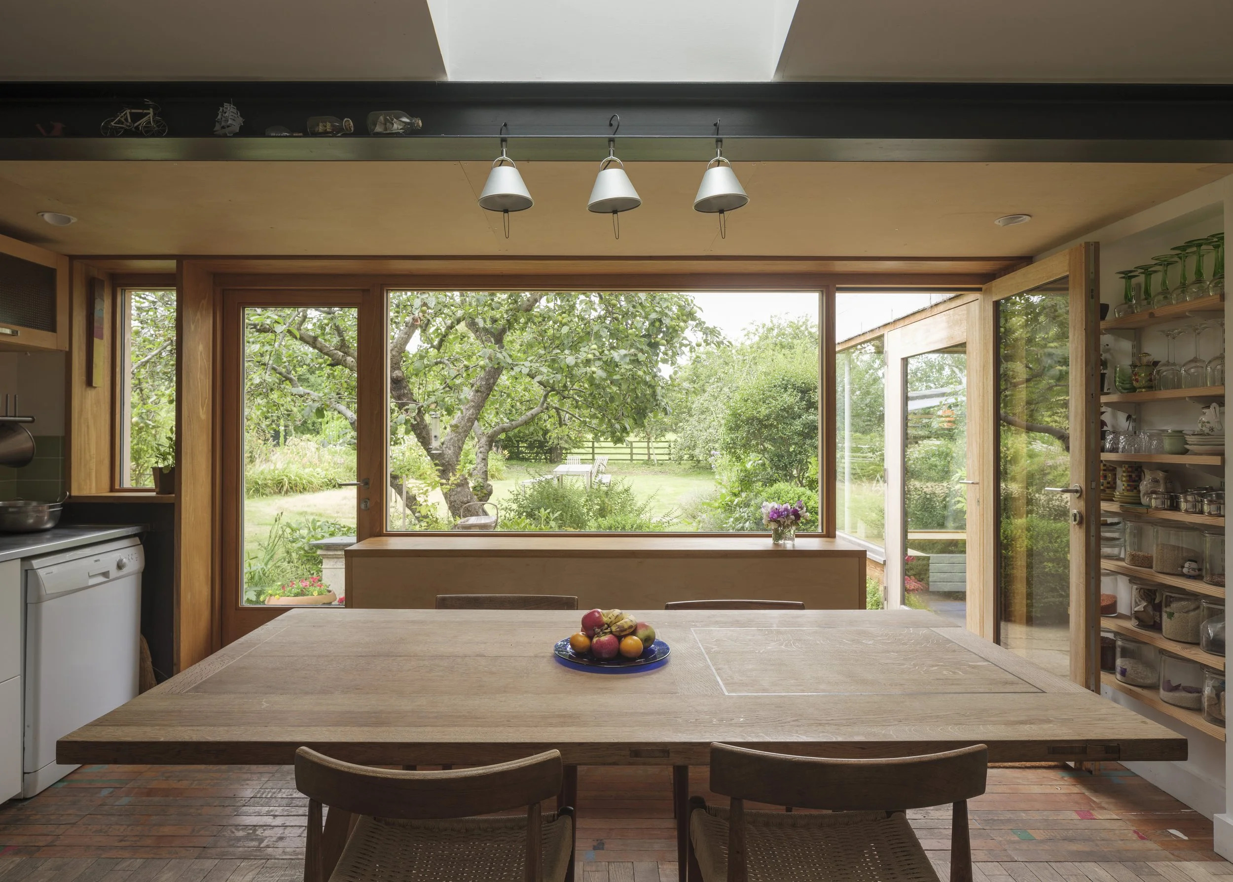 View of a bright kitchen with large windows overlooking a lush garden, a wooden dining table, and chairs. A fruit bowl with apples and oranges is on the table. The kitchen features shelves with glassware and a view outside showing trees and outdoor f