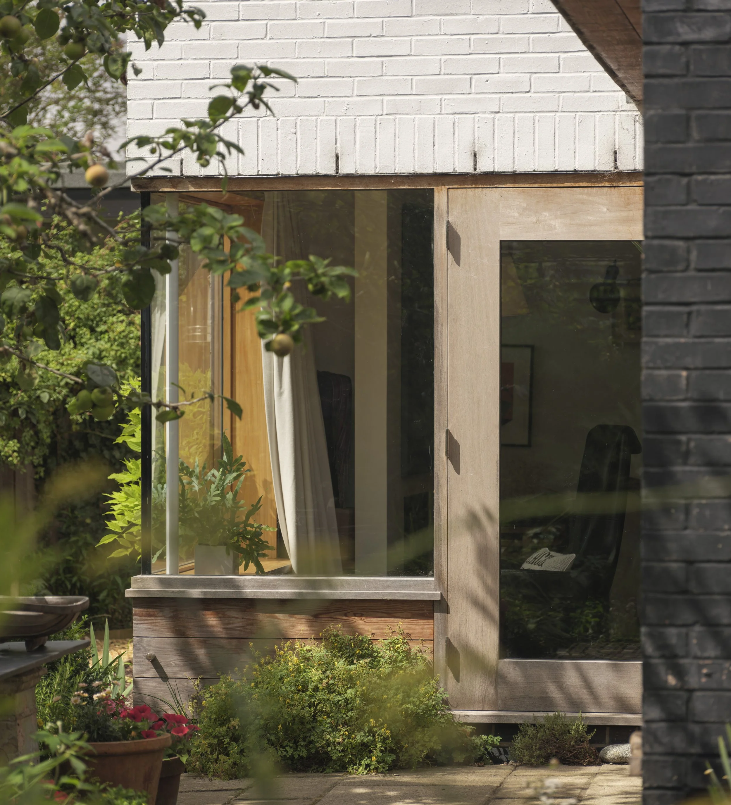 A close-up view of a garden with potted plants and flowering bushes in the foreground, and a modern house with large glass windows, wooden framing, and white brick walls in the background.