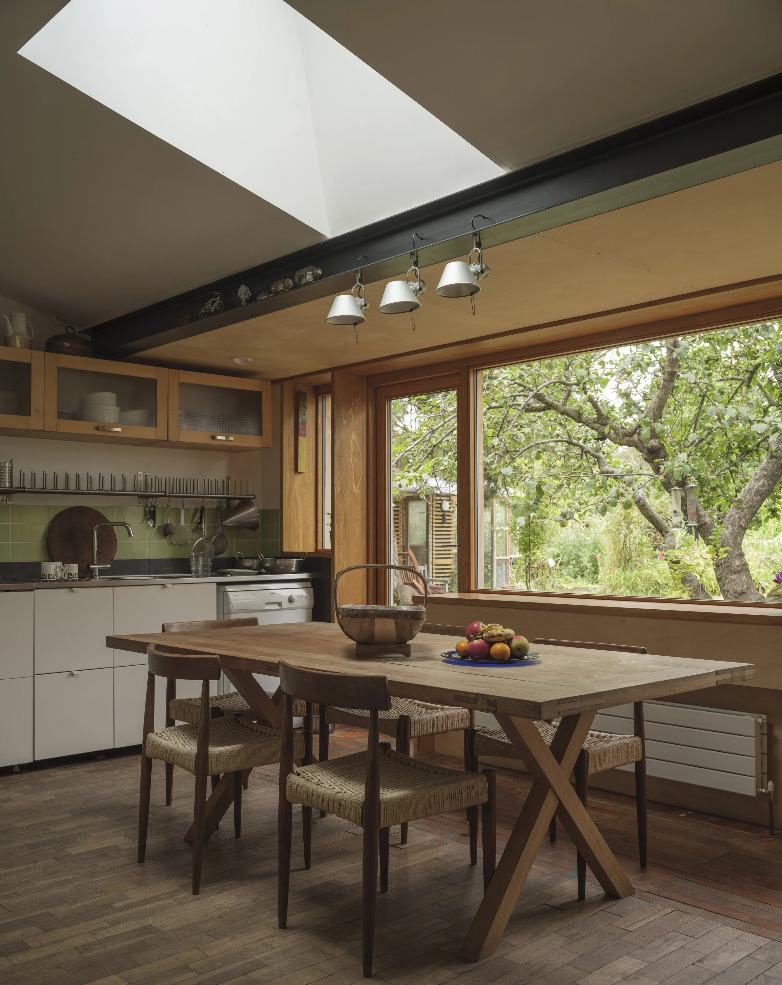 Kitchen with wooden table and chairs, large window with view of trees, skylight in ceiling, and hanging light fixtures.