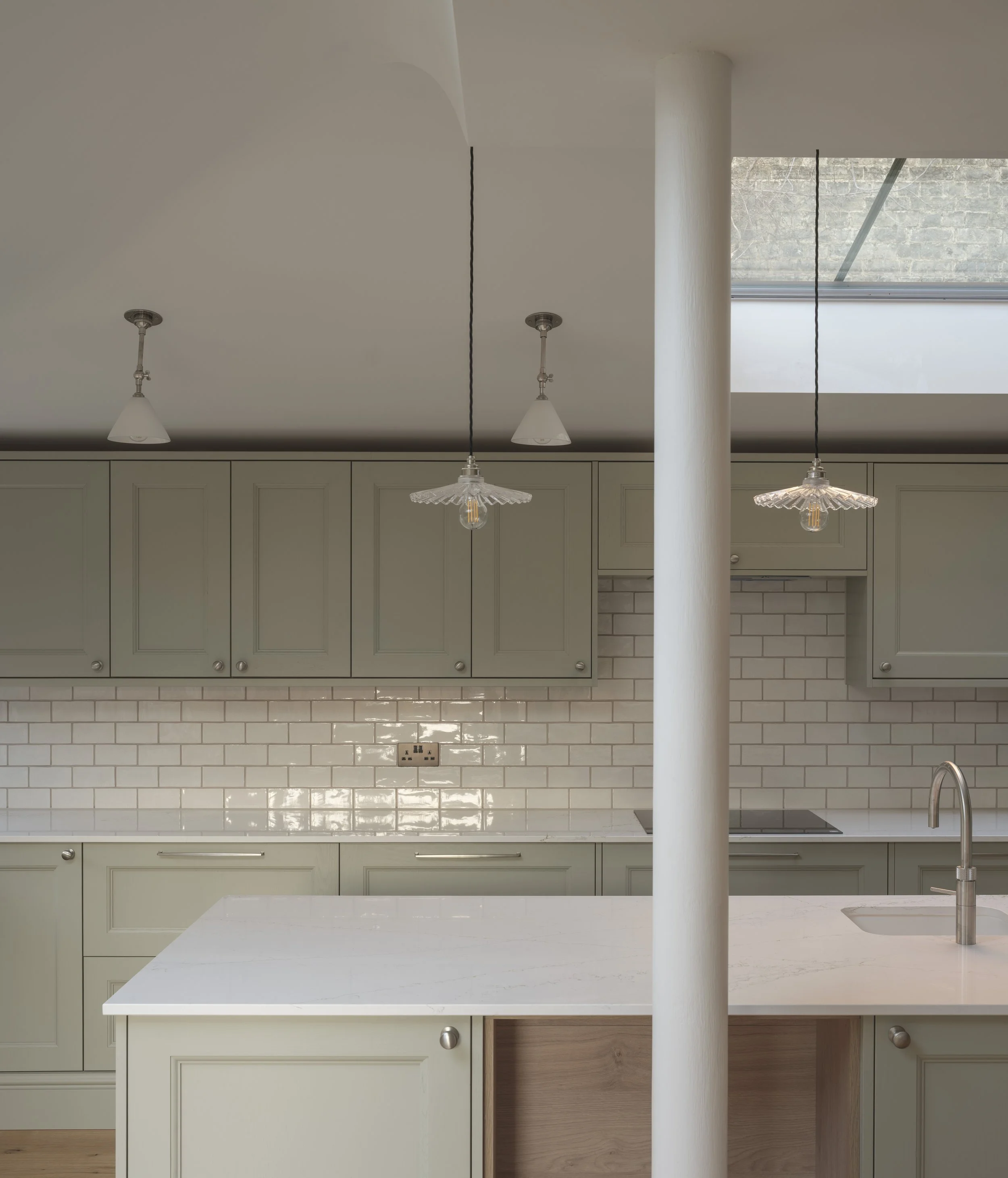 Modern kitchen with white cabinets, a white countertop, a white brick backsplash, a sink, a chrome faucet, and pendant lights hanging from the ceiling. There is a large window with a stone wall visible outside.