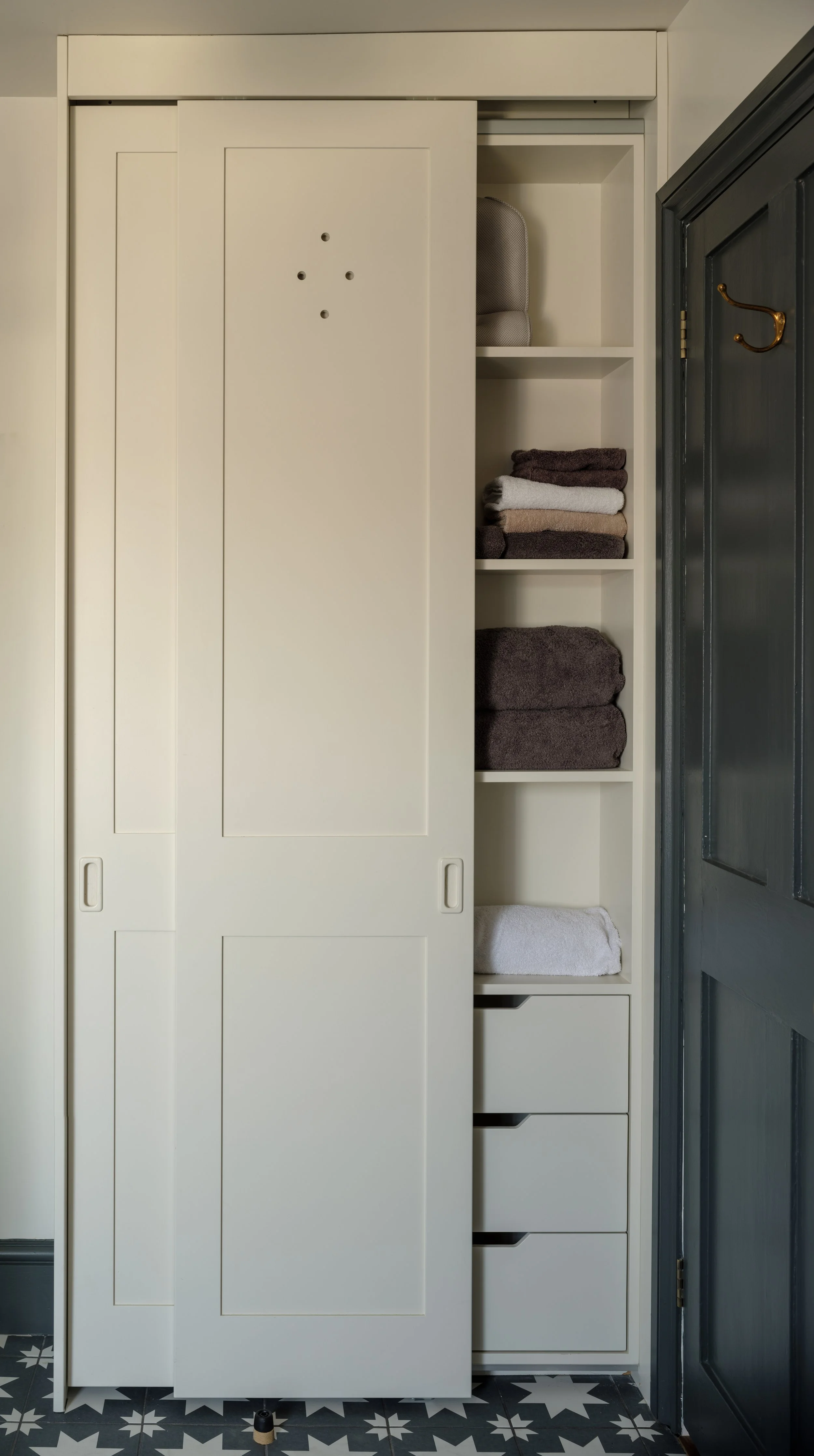 A white sliding storage closet with folded towels and a soft storage bin inside, situated next to a dark-colored door, on a black and white patterned tiled floor.