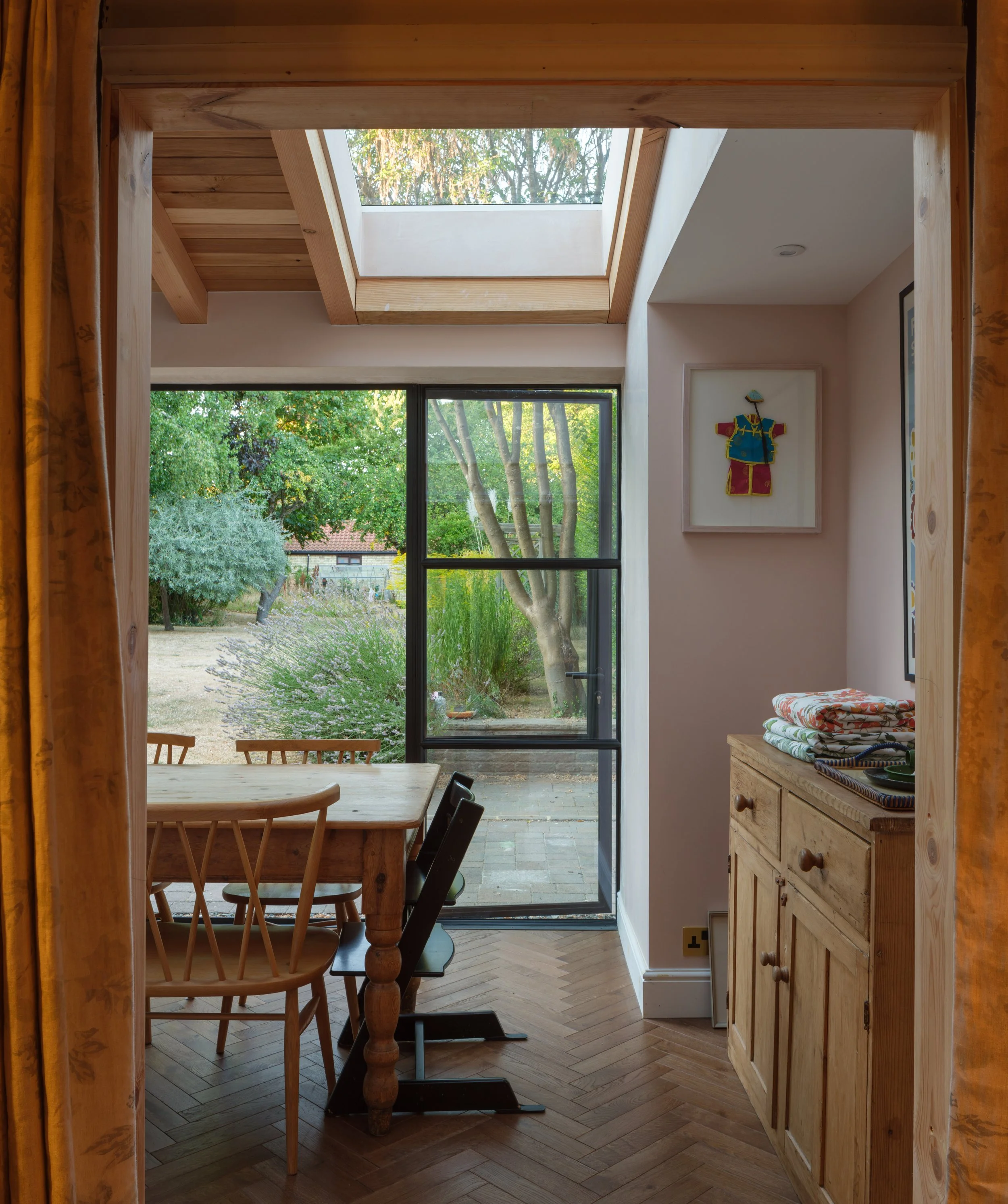 Interior view of a room with large windows showing a view of green trees outdoors. There is a wooden dining table with chairs, a wooden sideboard with folded towels, and framed artwork on the walls. A skylight is visible on the ceiling.