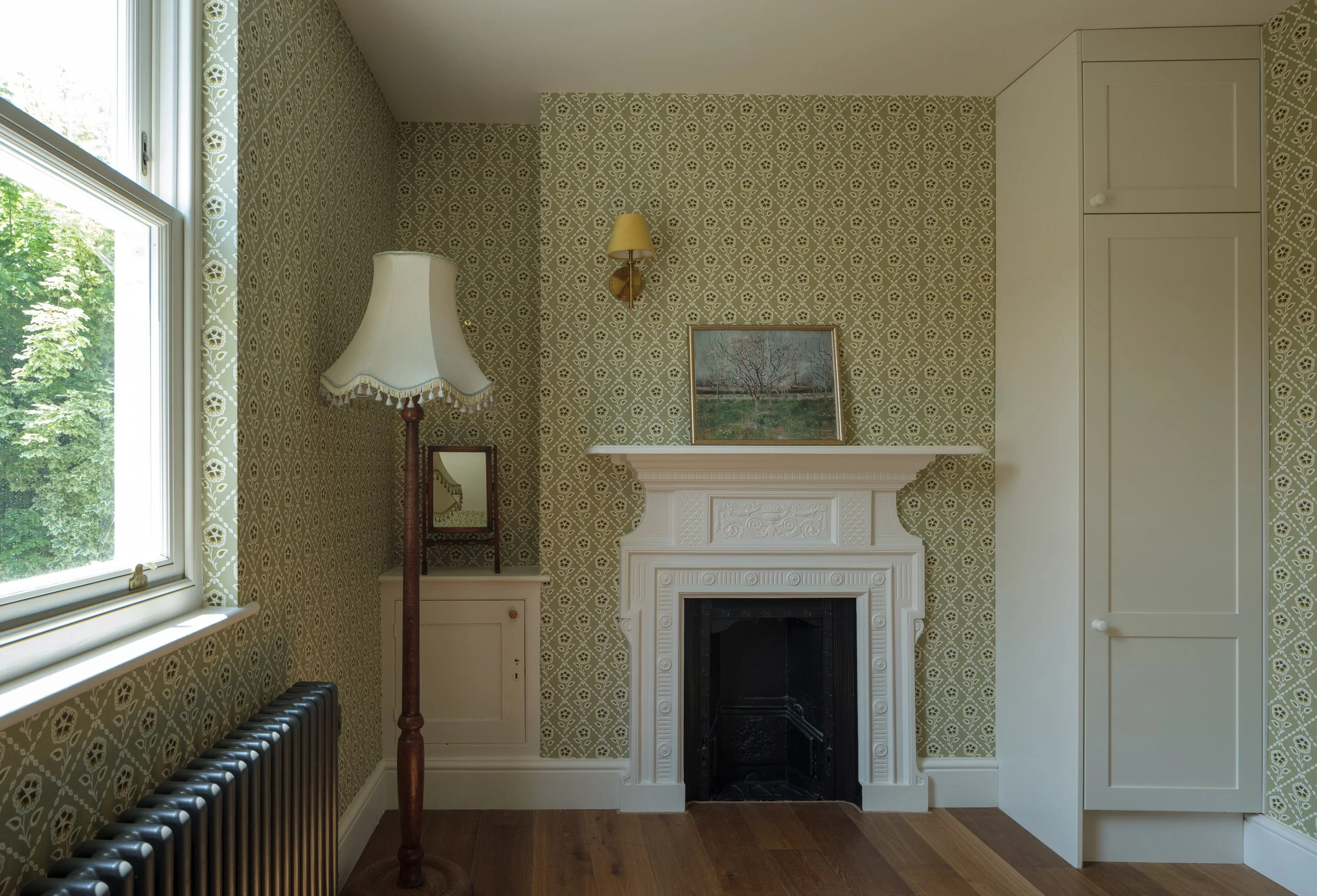 A cozy room with patterned wallpaper, a white fireplace, a framed painting on the mantel, a standing floor lamp, a small side table with a mirror, and a window with floral curtains showing green trees outside.