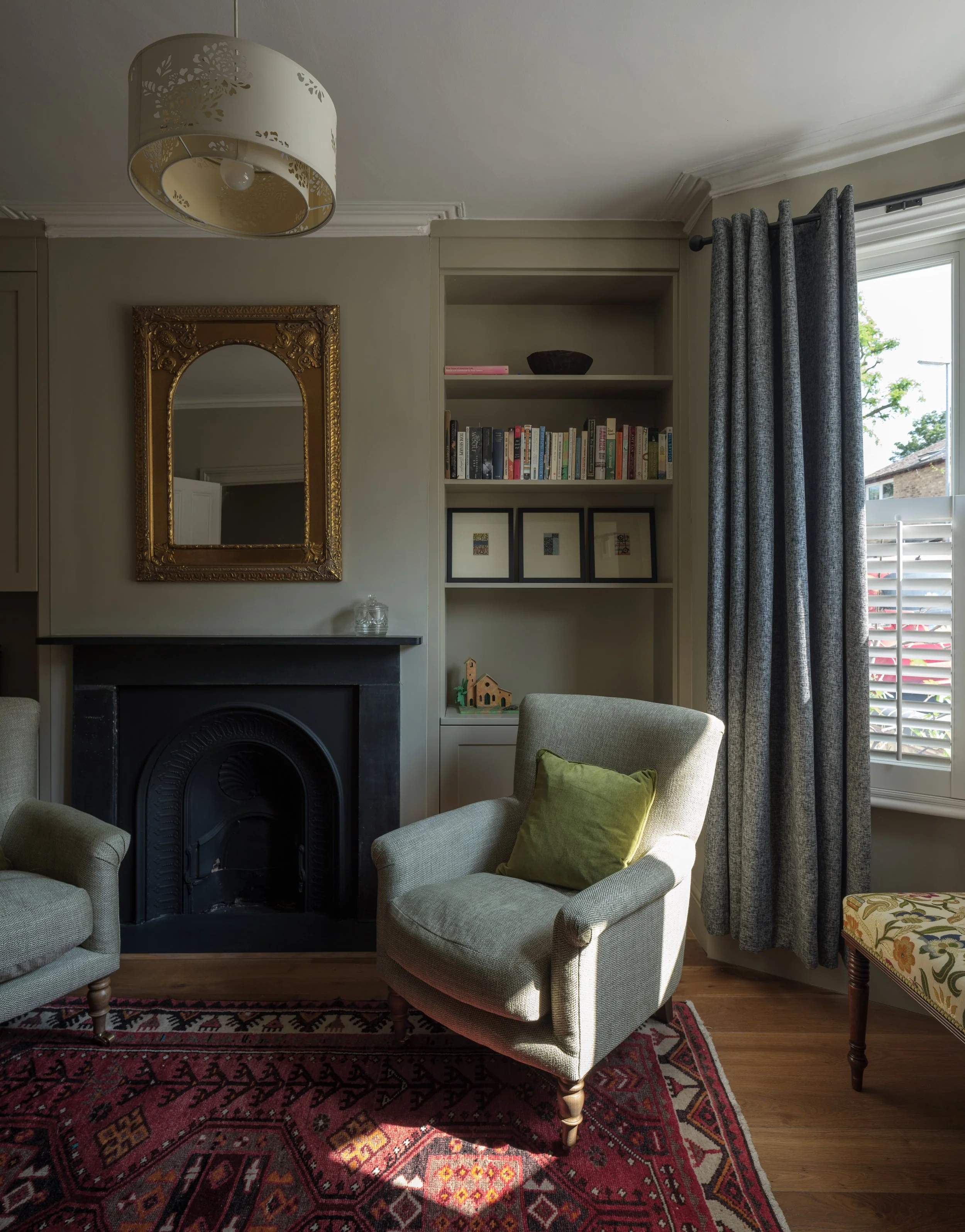A cozy living room corner with a beige armchair holding a green pillow, a fireplace with a black mantel, a large ornate gold-framed mirror on the wall, a white bookshelf with books and decor, a window with grey curtains, and a patterned red and black