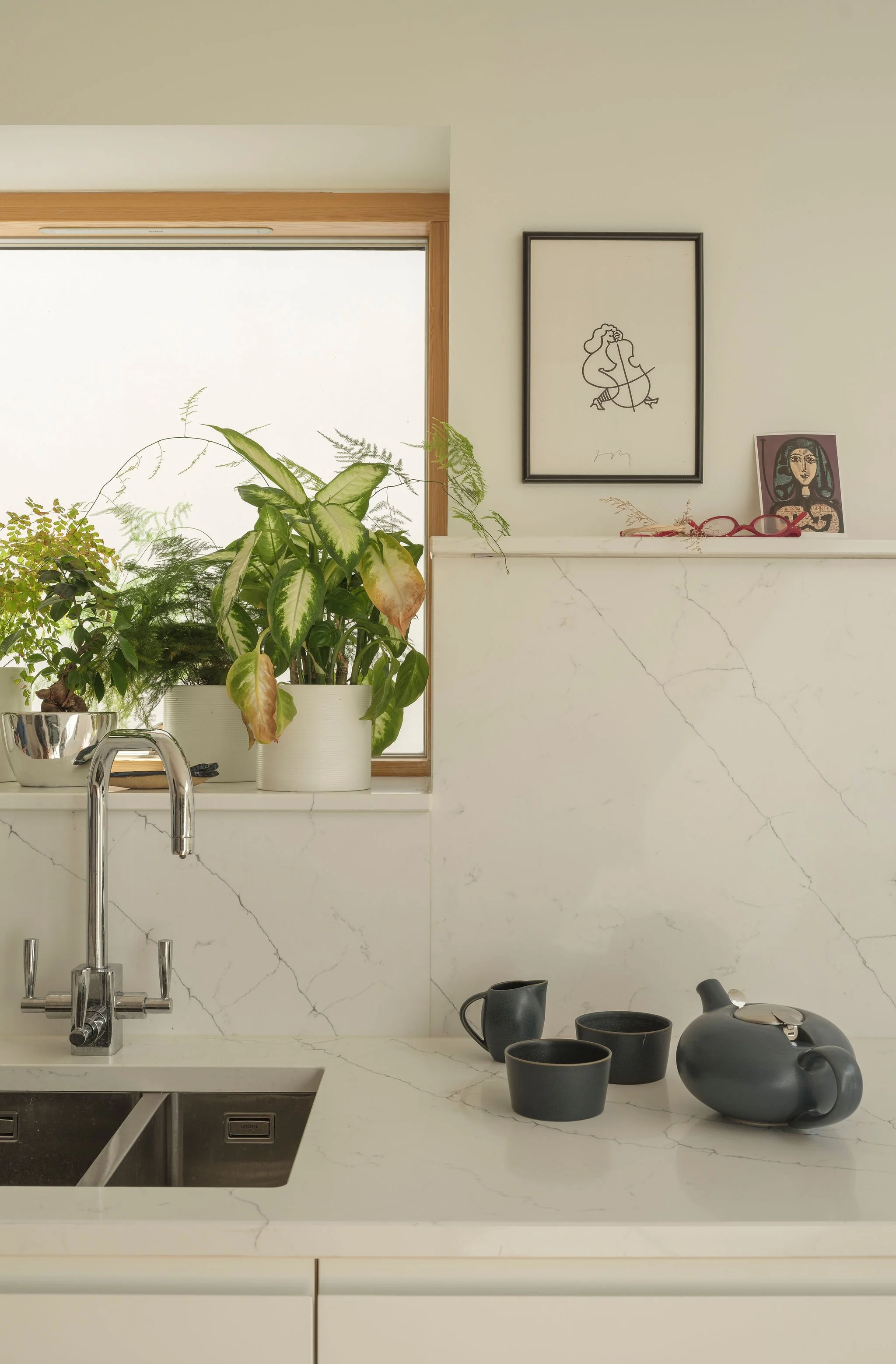 Modern kitchen countertop with a black teapot, cups, and a sink, decorated with houseplants, framed artwork, and kitchen utensils.
