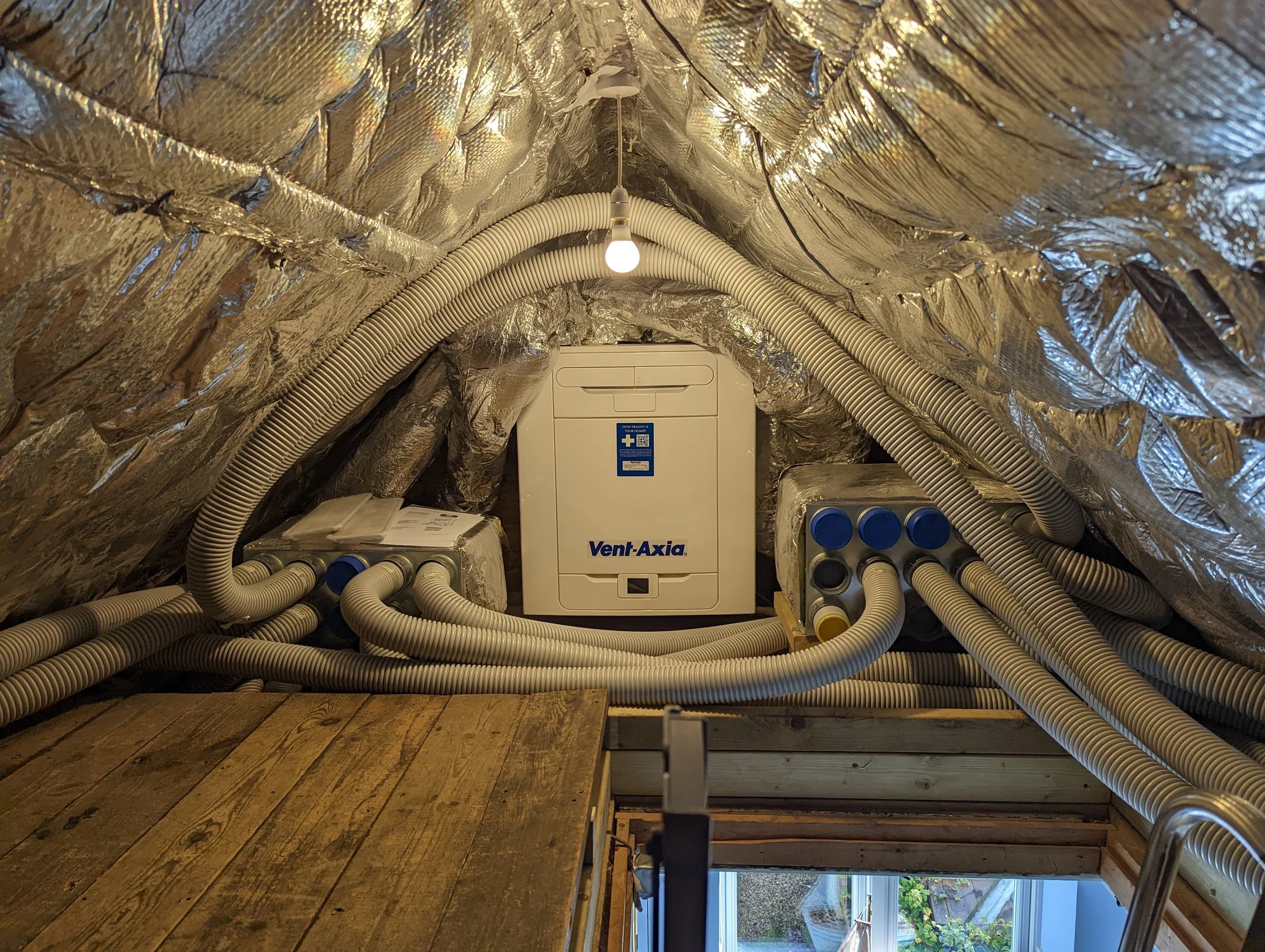 The image shows an attic space with reflective insulation on the walls and ceiling. There are flexible white ducts and a Vent-Axia ventilation unit, along with some boxes and wooden planks on the floor near a window.