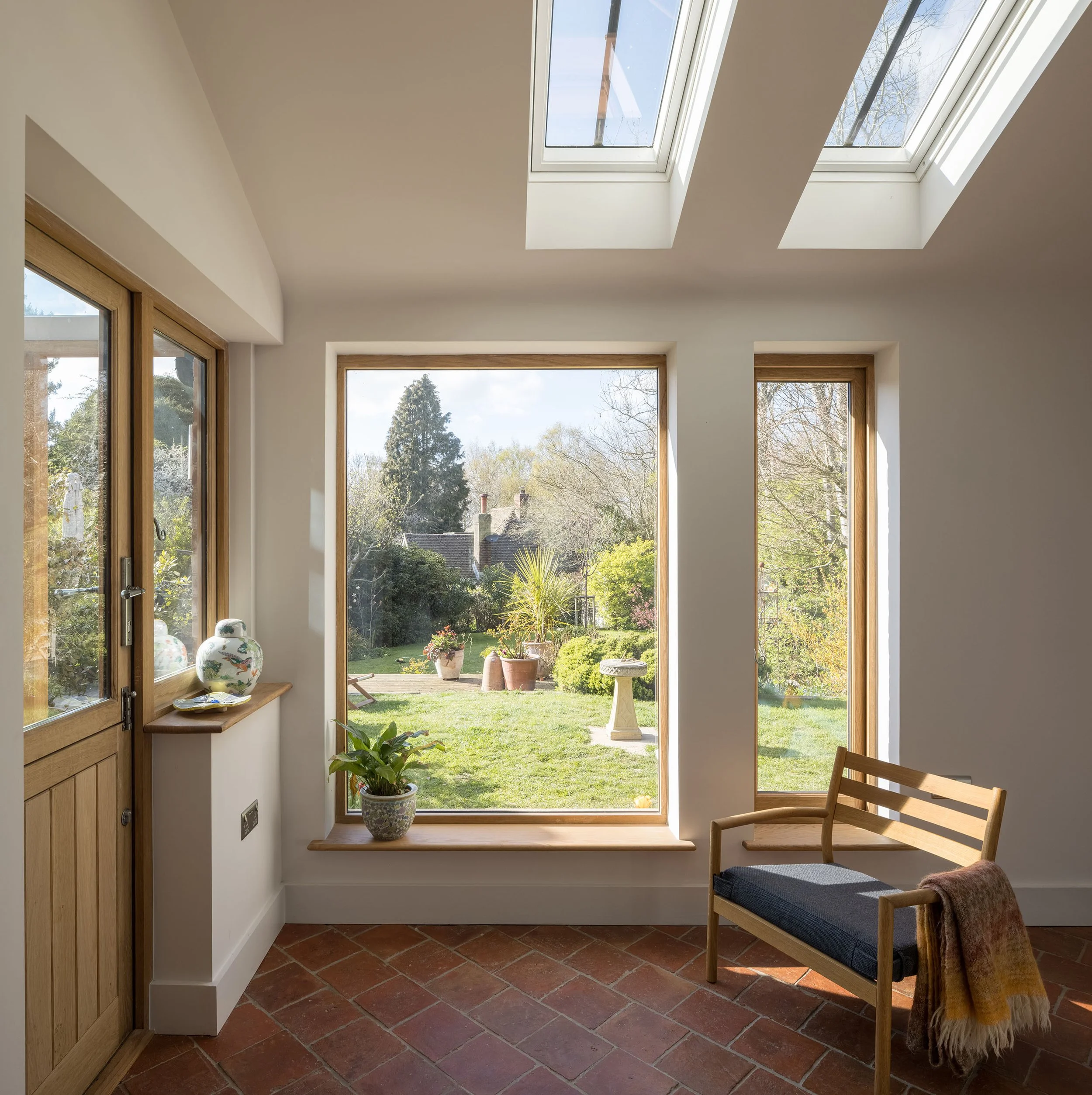 Bright room with large windows and skylights, wood frame window and door, potted plants, decorated vases, brick floor, outdoor garden visible through the windows, wooden chair with cushion and blanket.