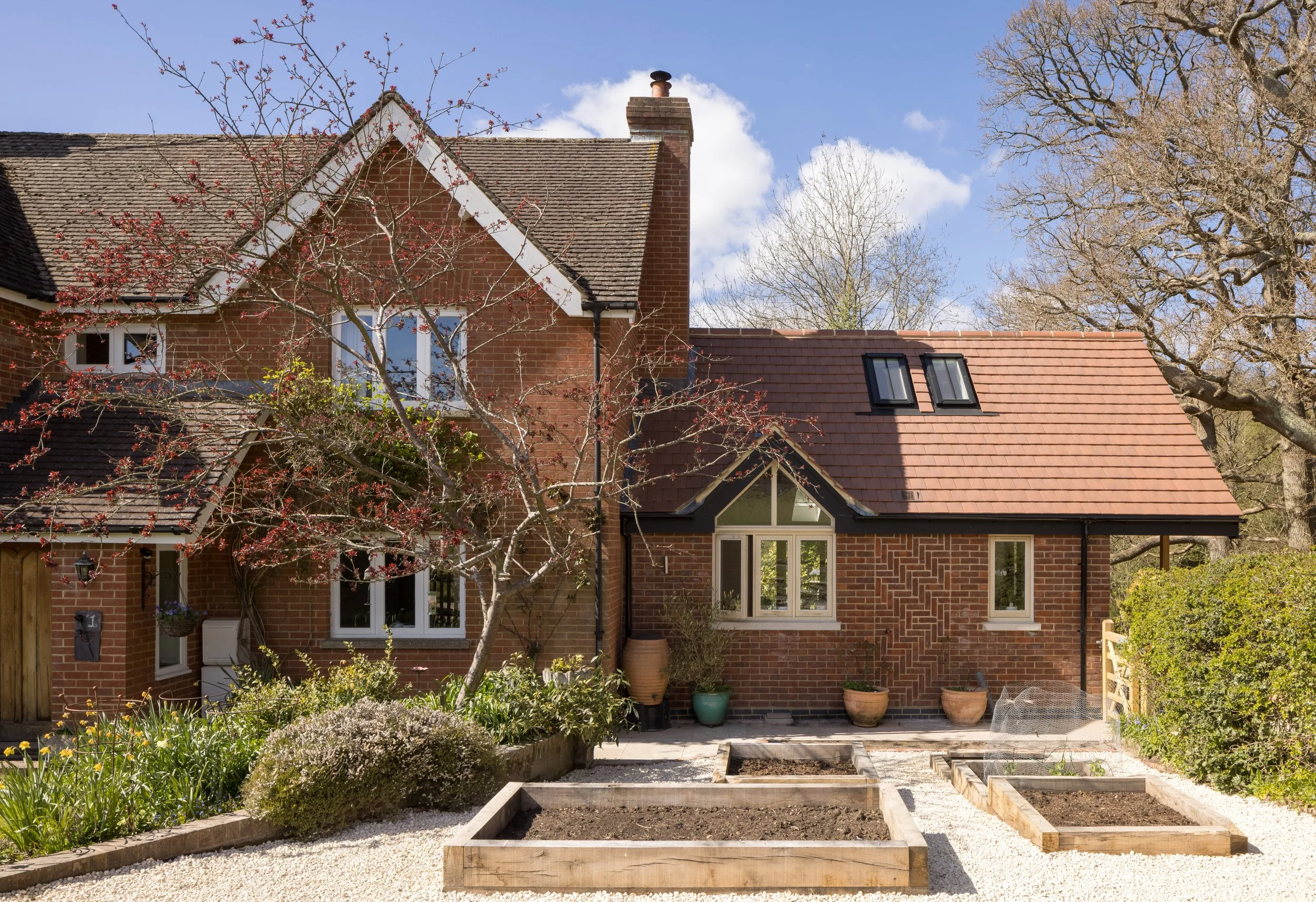 View of a backyard garden with raised vegetable beds and potted plants, brick house with dormer windows, trees, and a blue sky in the background.
