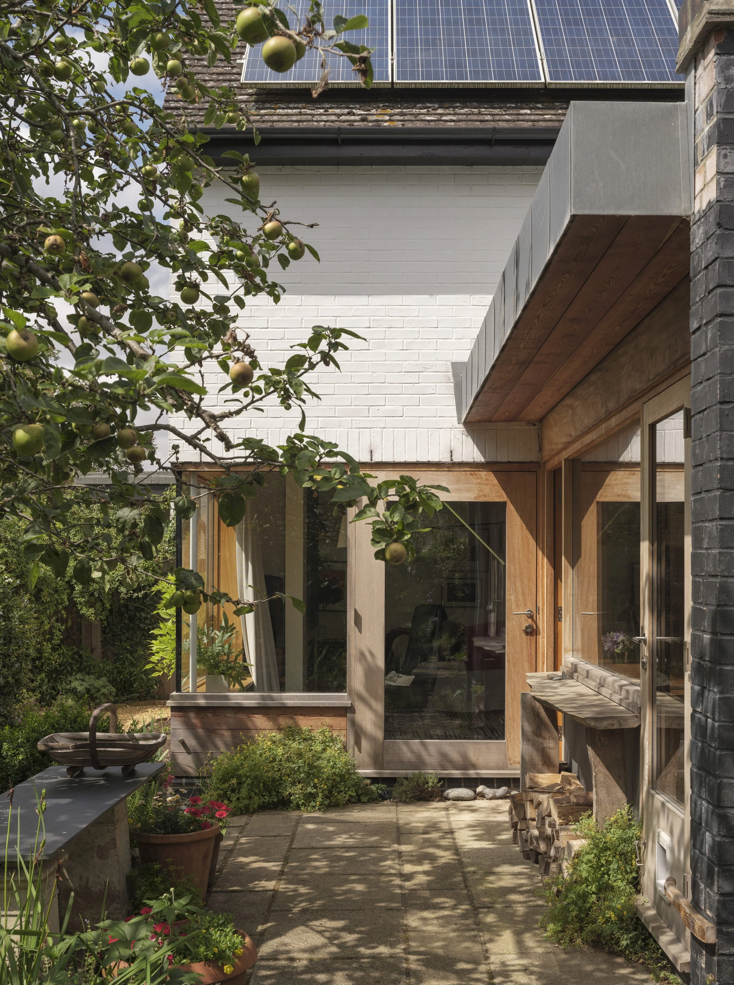 A garden courtyard view featuring a patio with potted plants, a tree with apples, a modern house with large windows, a white brick wall, solar panels on the roof, and a wooden overhang.
