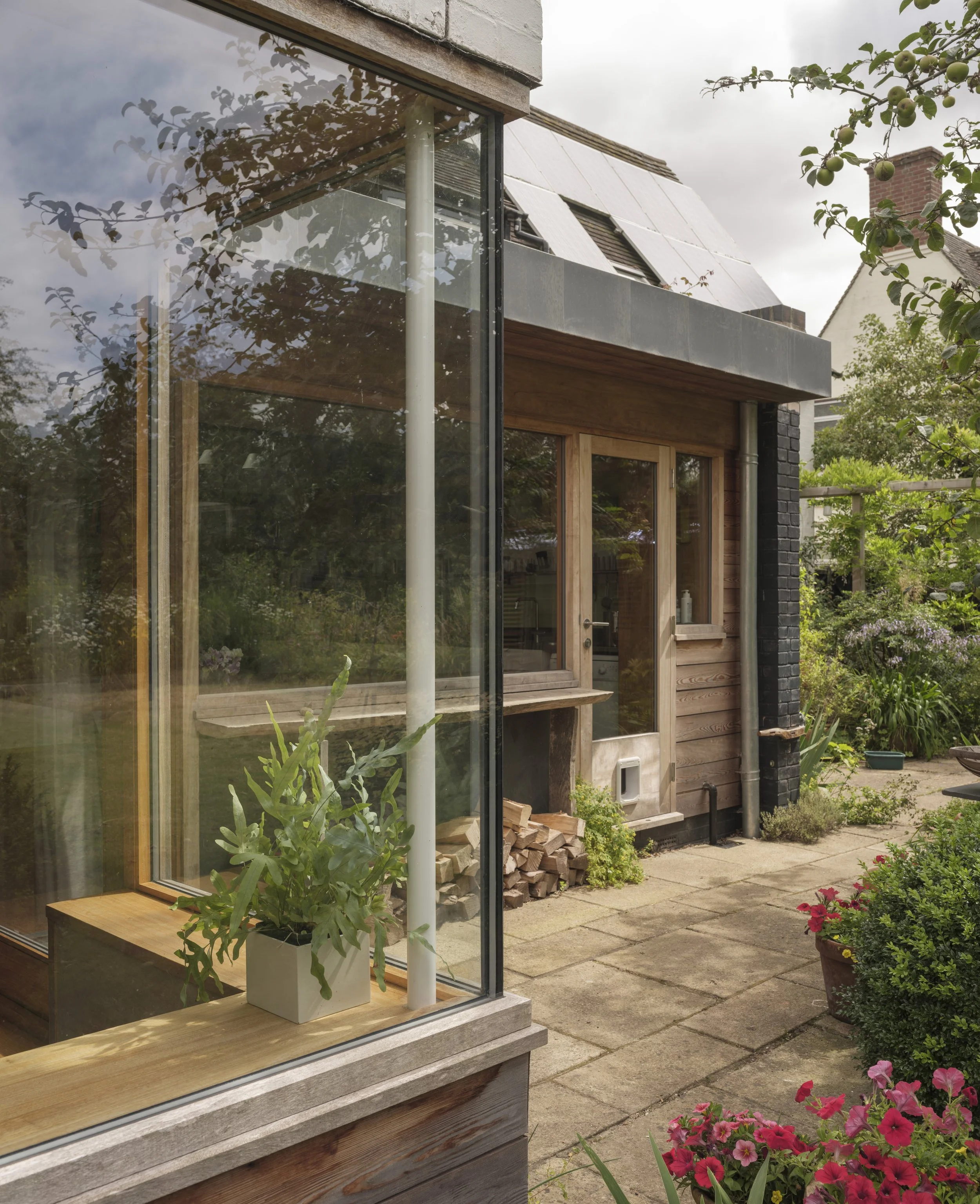 The image shows a view of a modern house's outdoor area. There is a large glass window with a wooden frame, and inside, a potted plant is visible on a wooden ledge. Outside, there are several plants, flowers in a pot, and a stack of wood near a black
