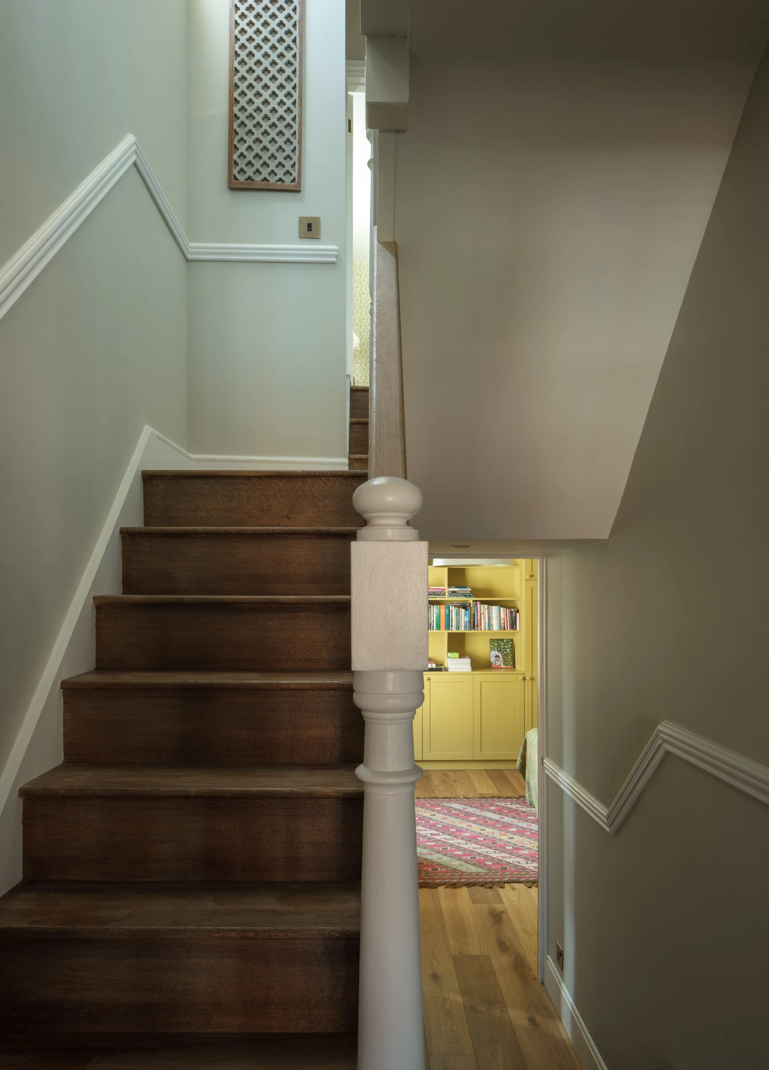 Interior view of a staircase with dark wooden steps and a white wooden banister. At the top of the stairs, there is a small hallway with an off-white wall, a decorative wall hanging, and a door leading to a room with a yellow bookshelf and patterned 