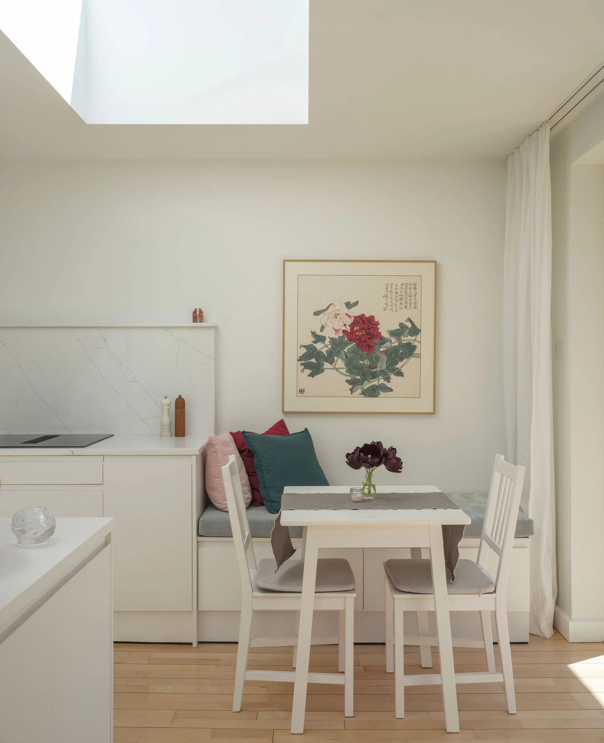 A bright, minimalist dining area with a white table and four white chairs, decorated with cushions in shades of green, beige, and pink. A small vase with dark purple flowers sits on the table. Behind the table, a built-in white cabinet with a marble-