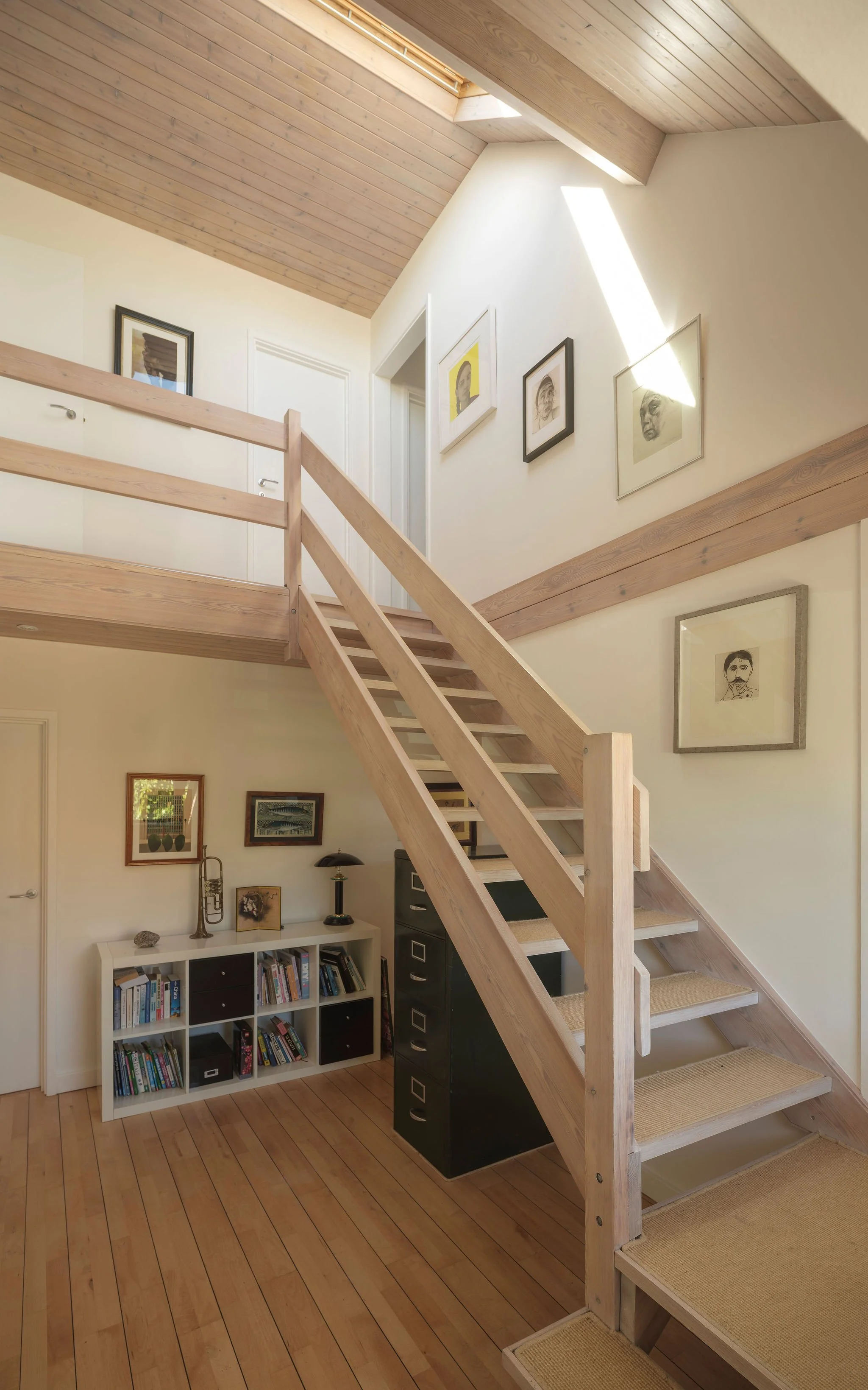 Interior view of a wooden staircase leading to a second-floor balcony with framed artwork on the walls and a bookshelf below. The ceiling has wooden paneling and skylights allowing natural light to illuminate the space.