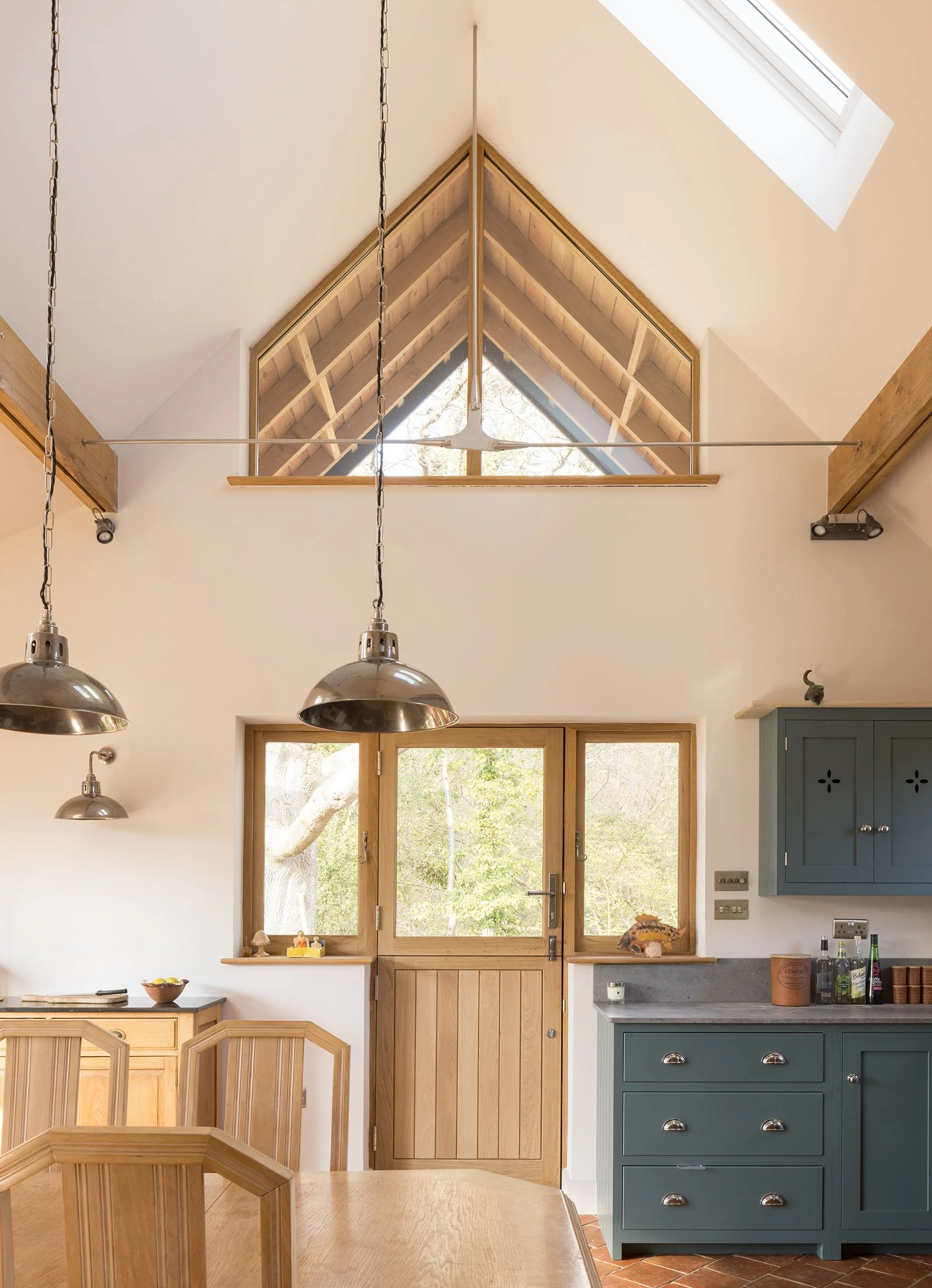 A kitchen with a high ceiling, a skylight, and a triangular window that shows trees outside. There are hanging pendant lights, a wooden dining table with matching chairs, and a blue cabinet with drawers and a countertop.