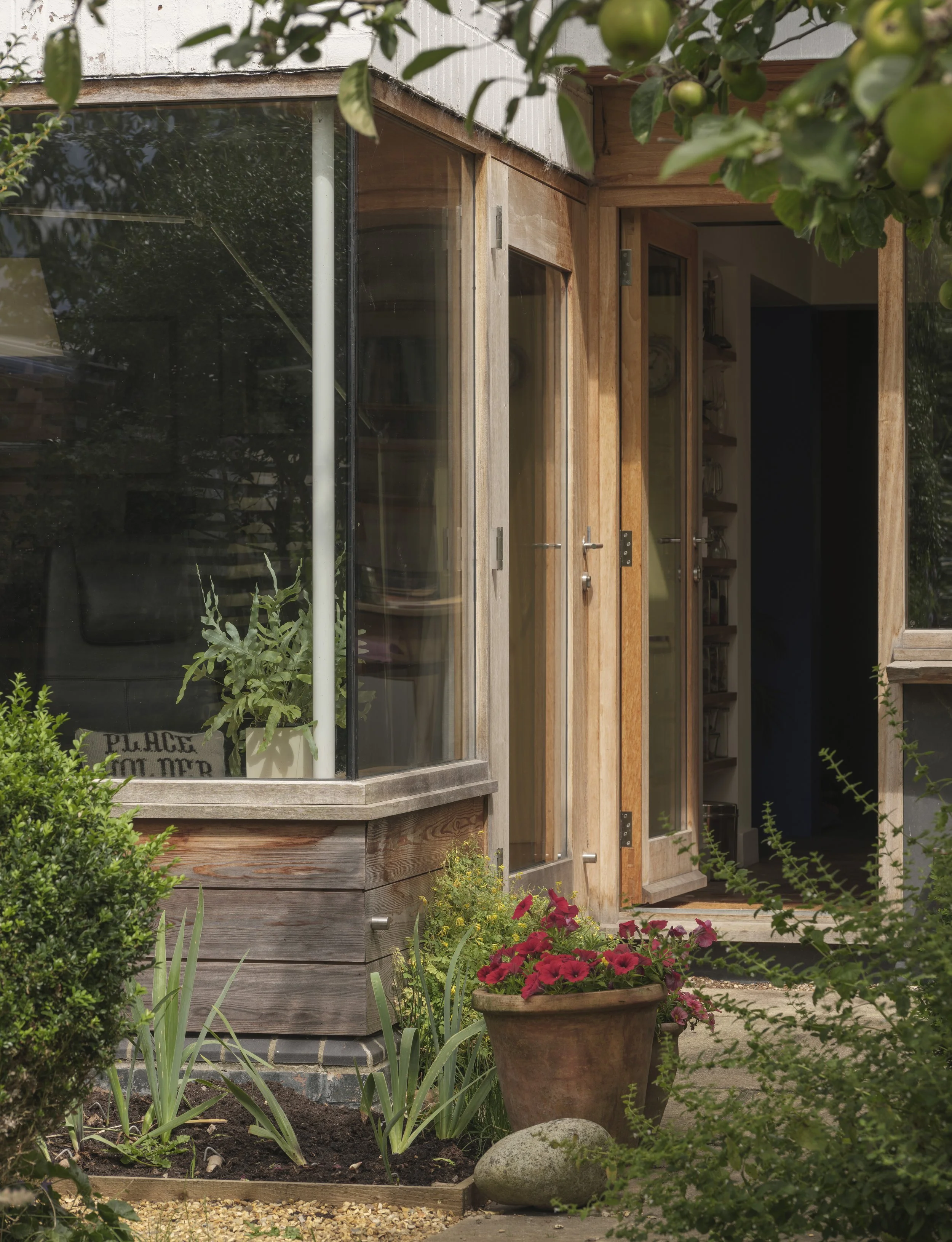 View of a house with a wooden and glass exterior, open door revealing interior shelves, surrounded by a garden with various plants, flowers, a large pot of red flowers, and garden decor.