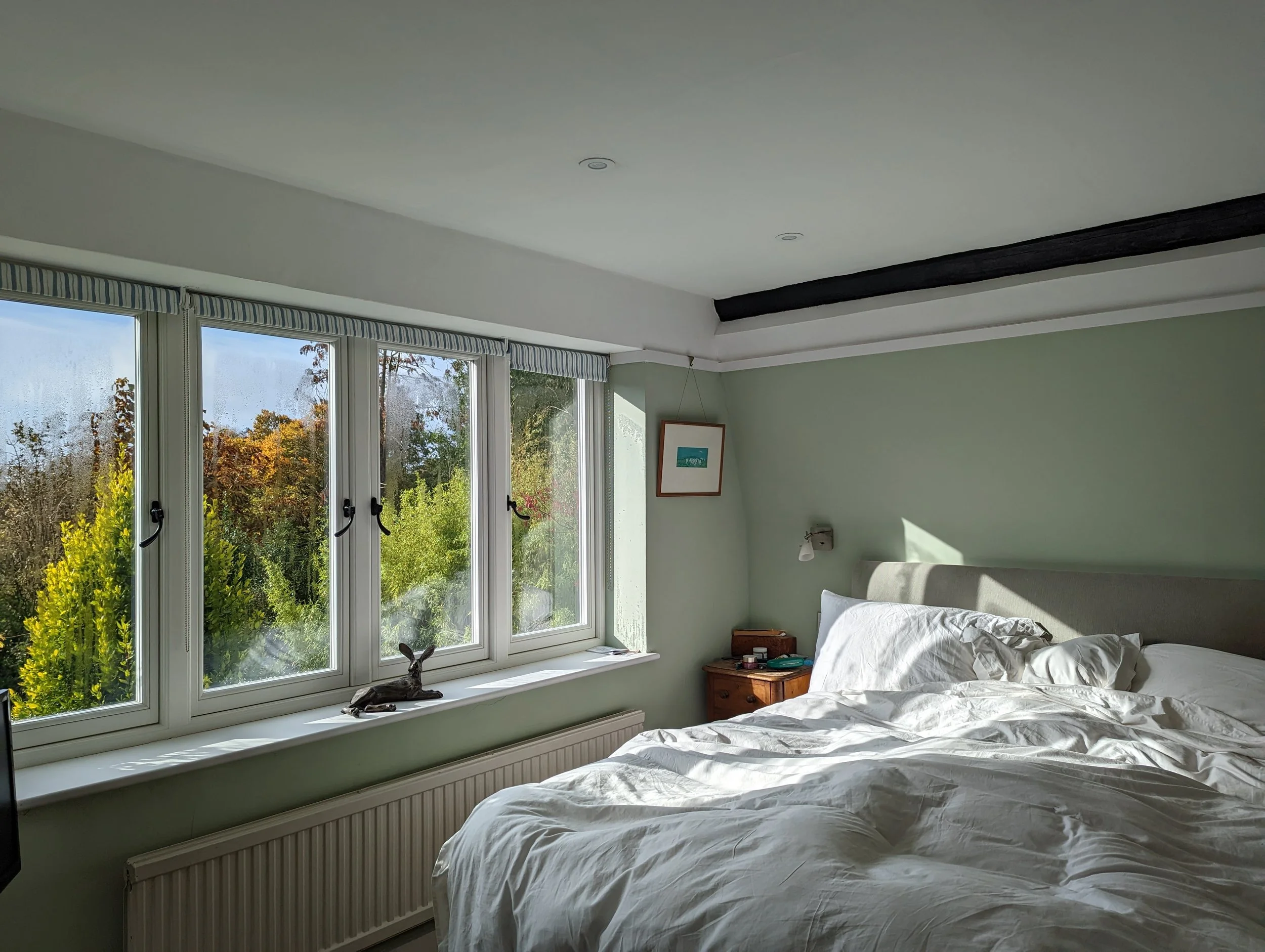 A bedroom with a large window showing trees outside, a bed with disheveled white bedding, a wooden nightstand with items, and a framed picture on the wall.