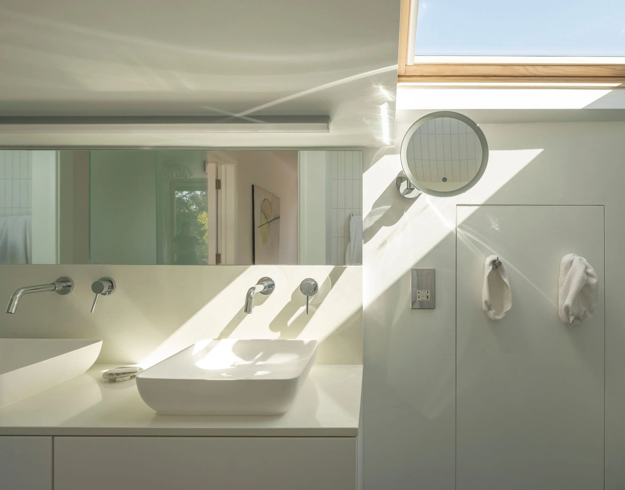 A modern bathroom with a white marble countertop, two wall-mounted sinks with chrome faucets, a large mirror, and a skylight letting in natural light.