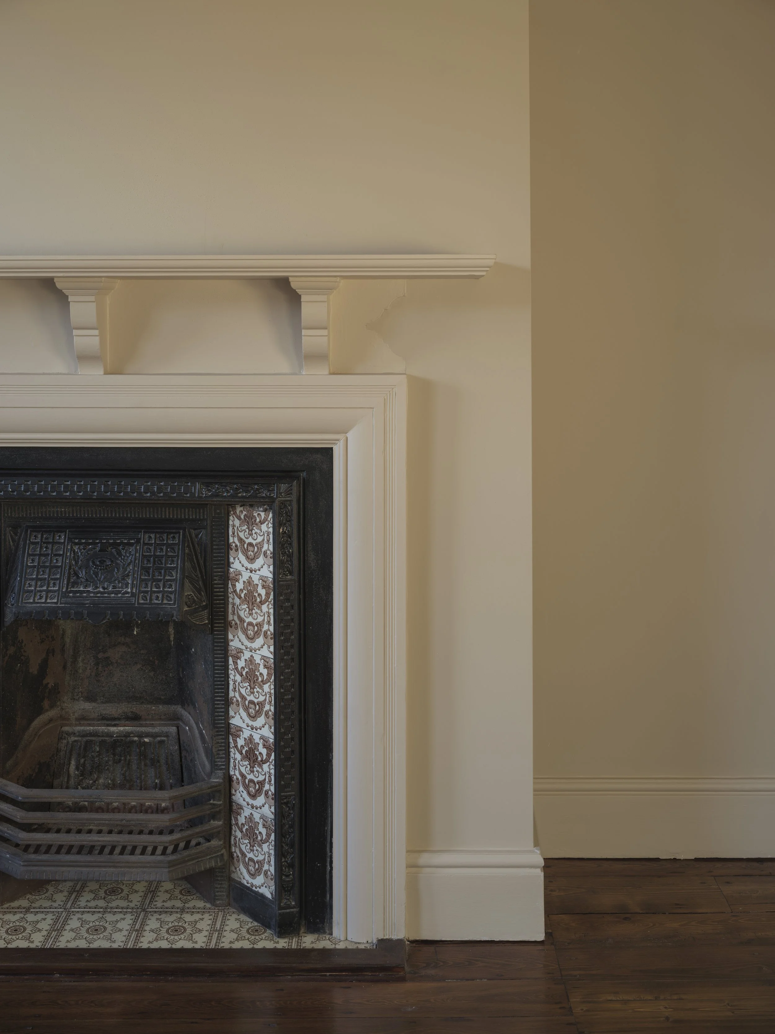Part of a fireplace with ornate black metal fire grate, surrounded by cream-colored trim, on a vintage tile hearth, adjacent to a wall with crown molding, hardwood floor.