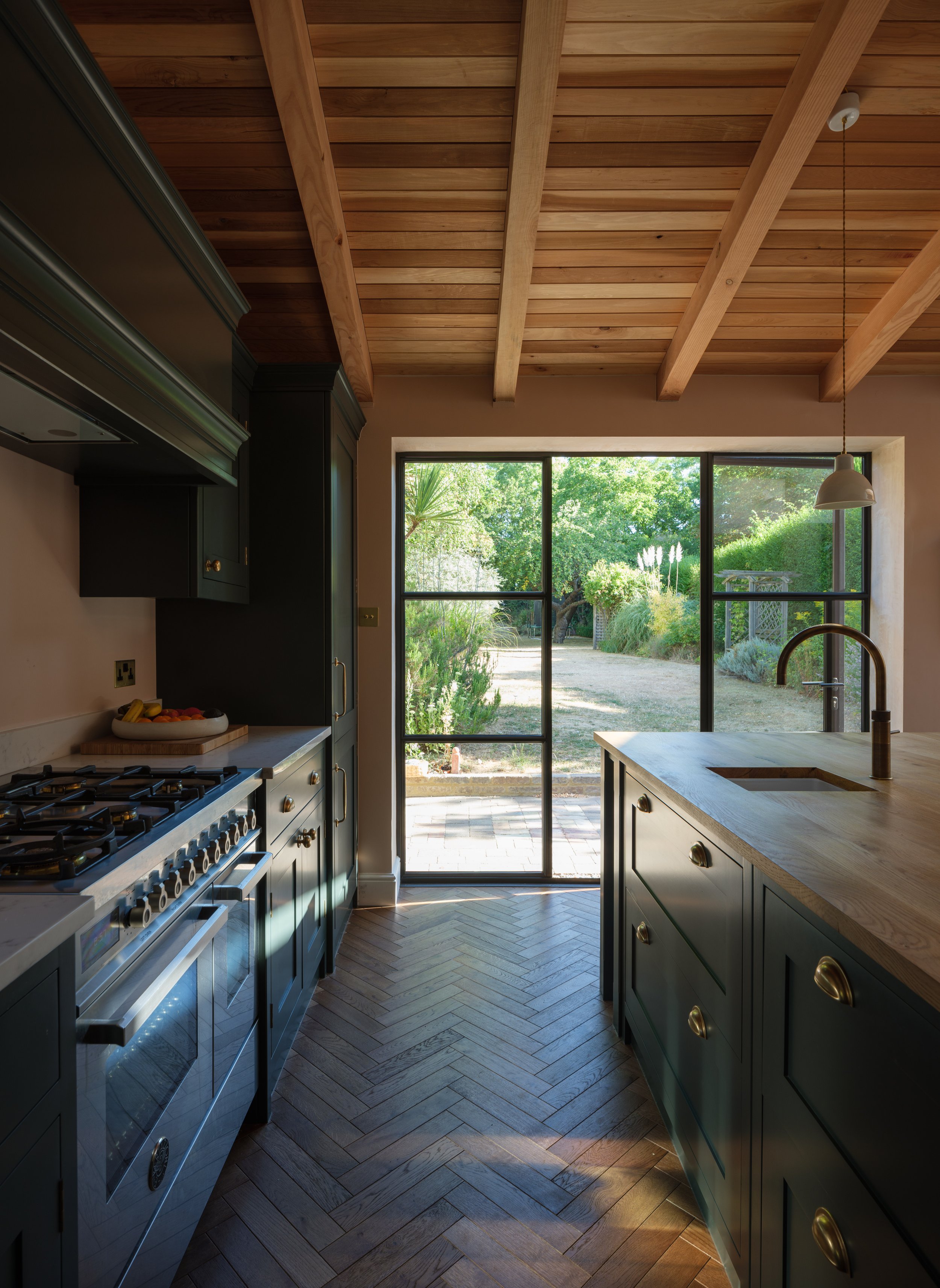 A modern kitchen with a wooden ceiling, dark green cabinets, stainless steel appliances, and large sliding glass doors leading to a lush outdoor garden.