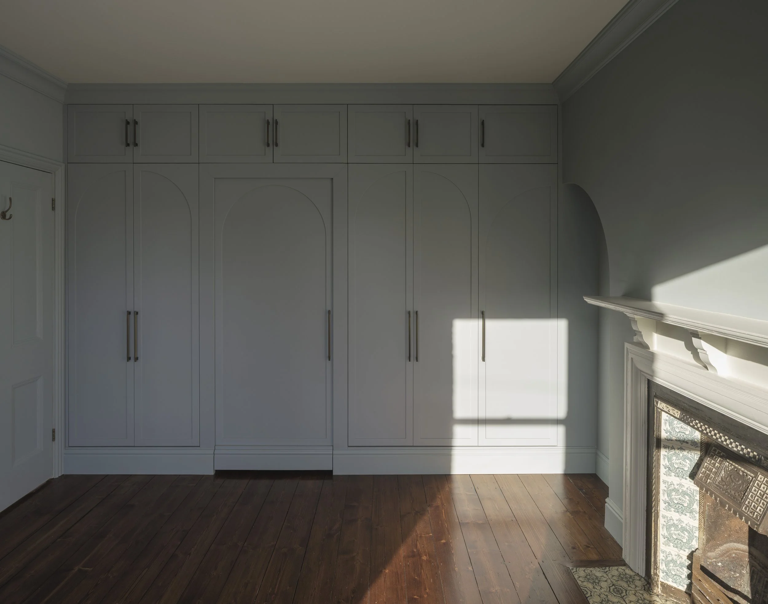 White built-in cabinets with black handles in a room with hardwood floor and a white fireplace mantel. Sunlight casts a shadow on the wall.