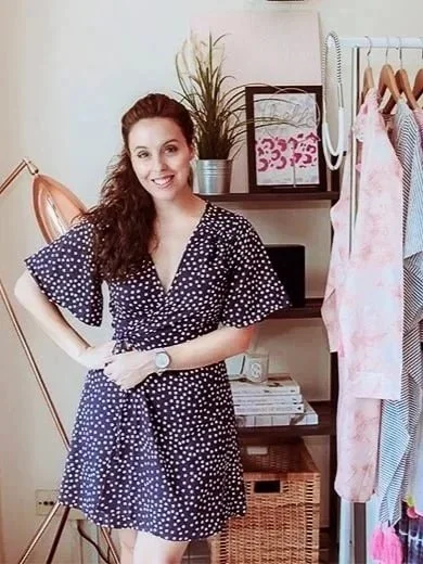 A woman standing inside a room with clothing and decorative items. She is wearing a navy blue dress with white polka dots, smiling, and has long, curly brown hair.