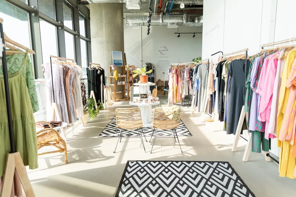 Interior of a clothing boutique with racks of colorful dresses and garments, a seating area with two wicker chairs, and large windows allowing natural light.