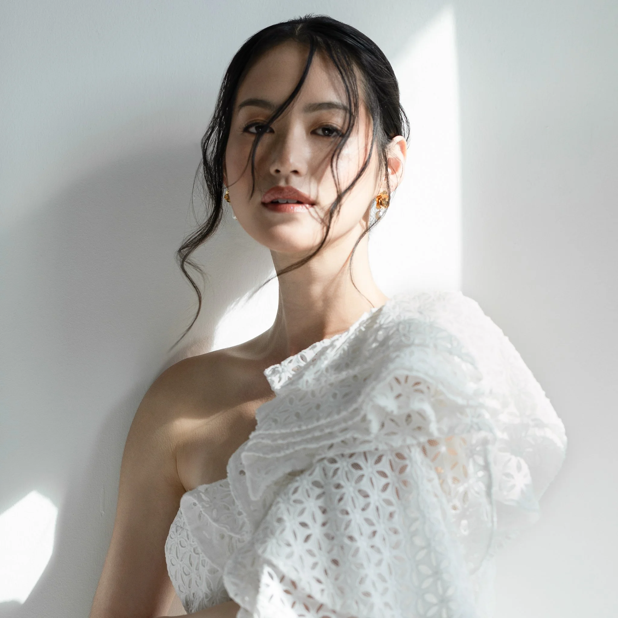 A woman with dark, slightly curly hair and gold earrings wearing a white, eyelet-patterned off-the-shoulder dress, standing against a white wall with sunlight creating shadows.