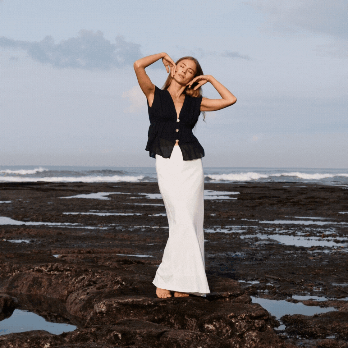 A woman with long hair in a black sleeveless top and white long skirt standing on rocks at the beach during daytime.