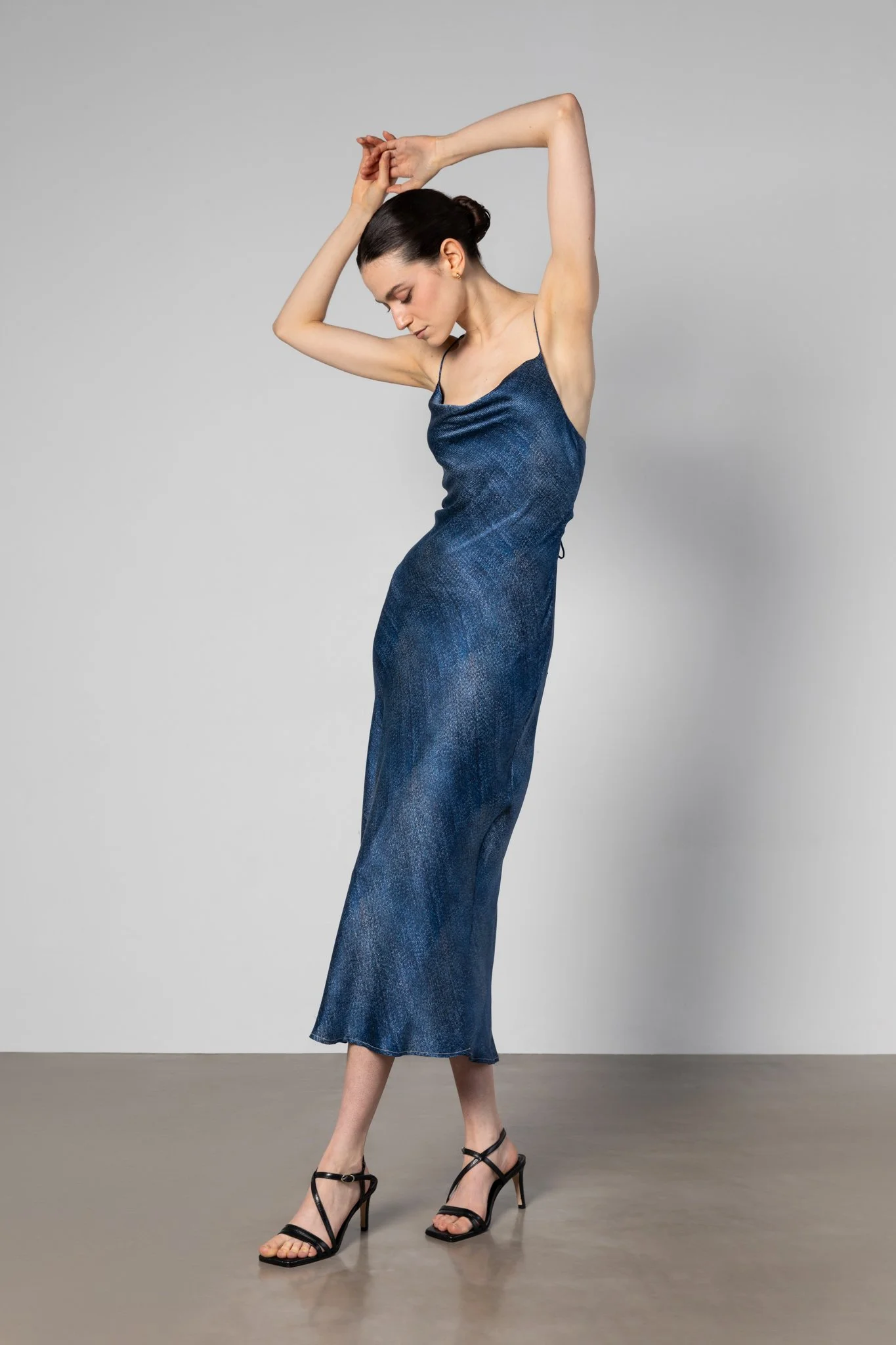 A woman in a blue slip dress and black high-heeled sandals stands against a plain background, her arms raised above her head, with her eyes closed.
