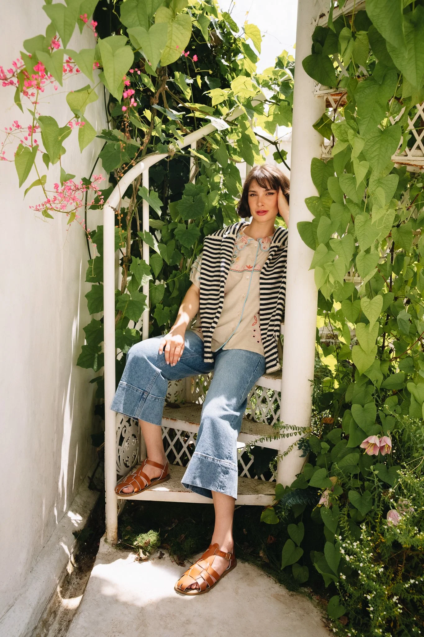A woman sitting on a white, spiral outdoor staircase surrounded by green leafy plants and pink flowers, with sunlight shining on her.