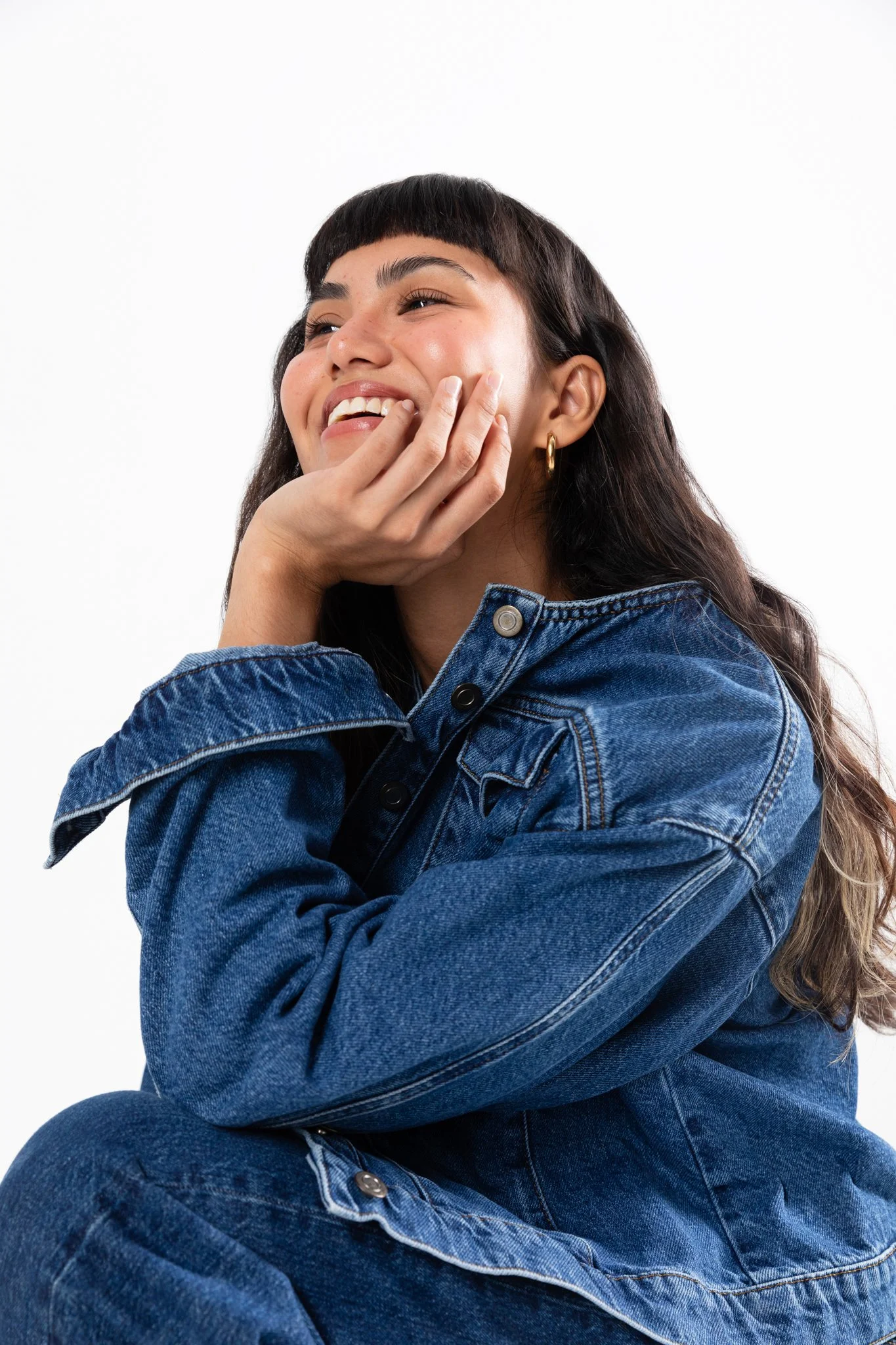 A young woman with dark brown hair, wearing a denim jacket, smiling and resting her chin on her hand, against a plain white background.