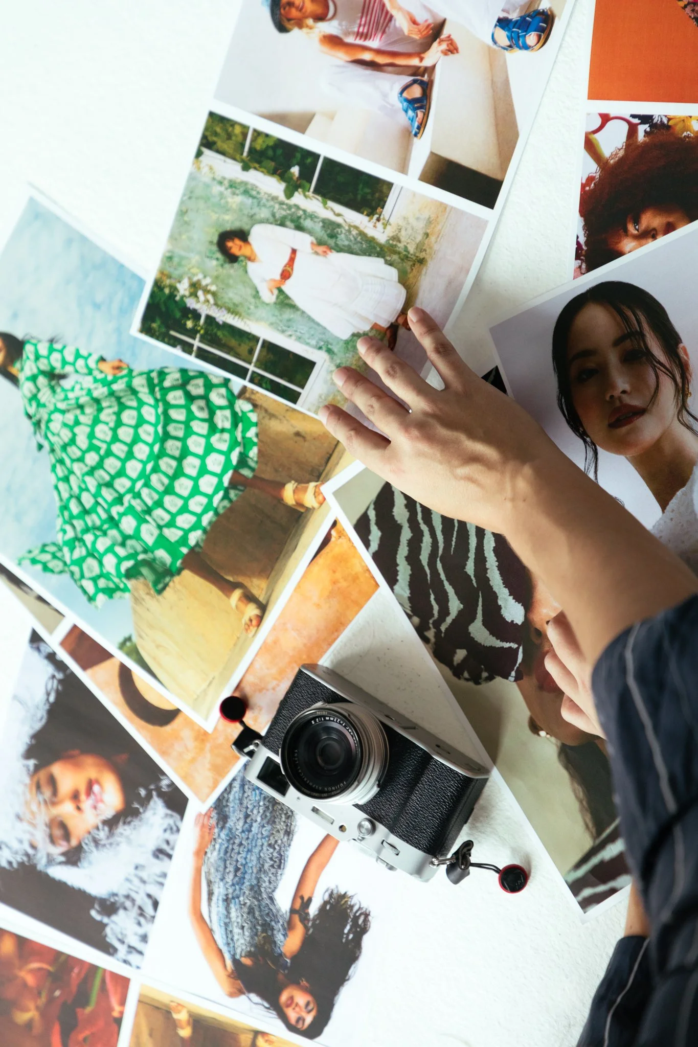 Person examining printed photos of women in various outfits and settings on a white table with a camera nearby.