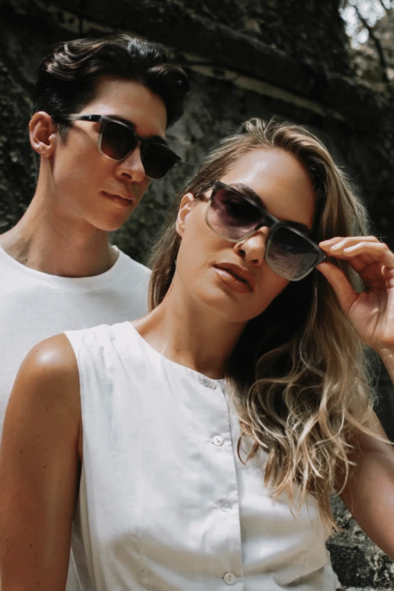 A man and woman wearing sunglasses outdoors, with the man standing behind the woman, both in casual white clothing.