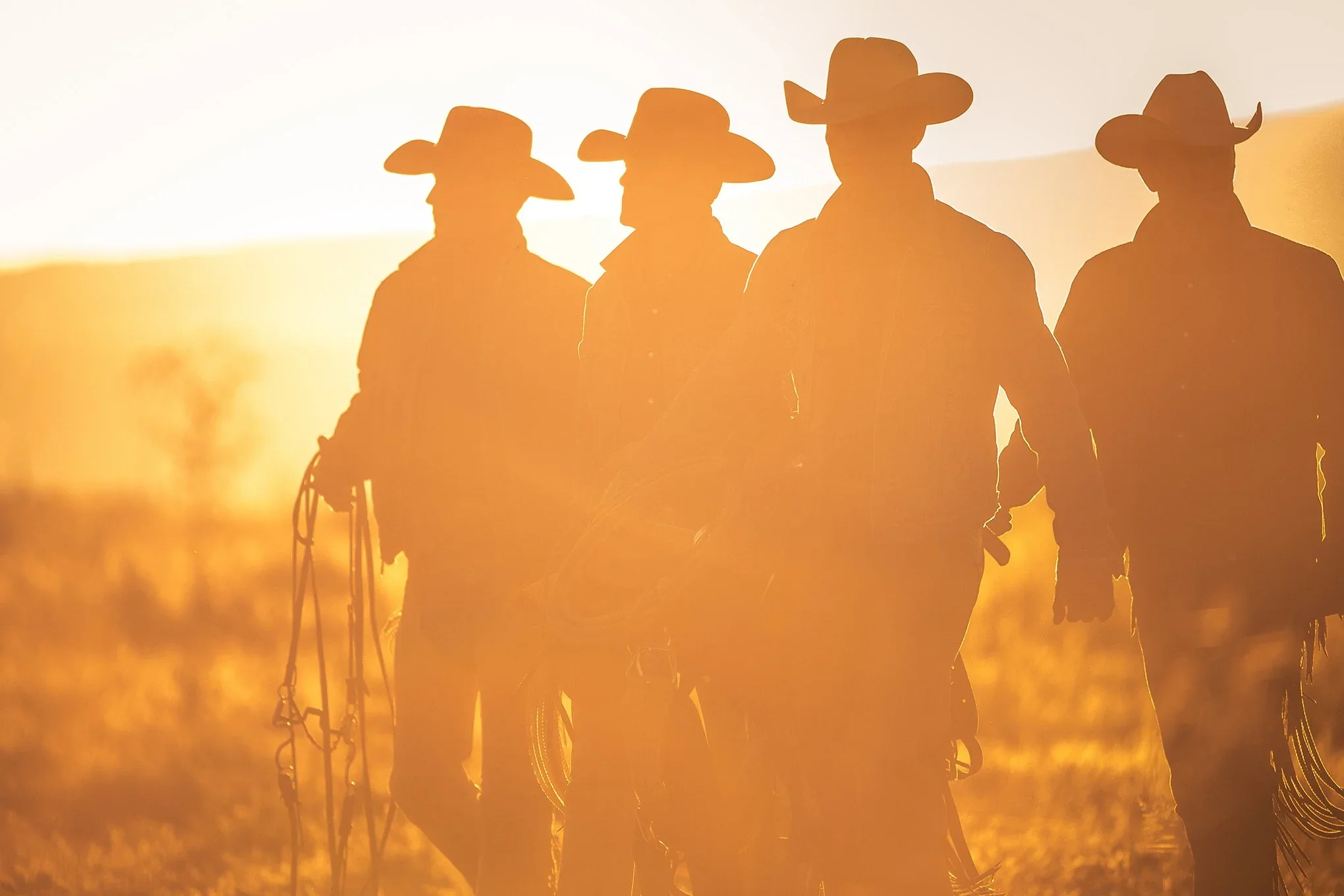 Silhouetted cowboys walking through golden dust at sunset during a cattle drive