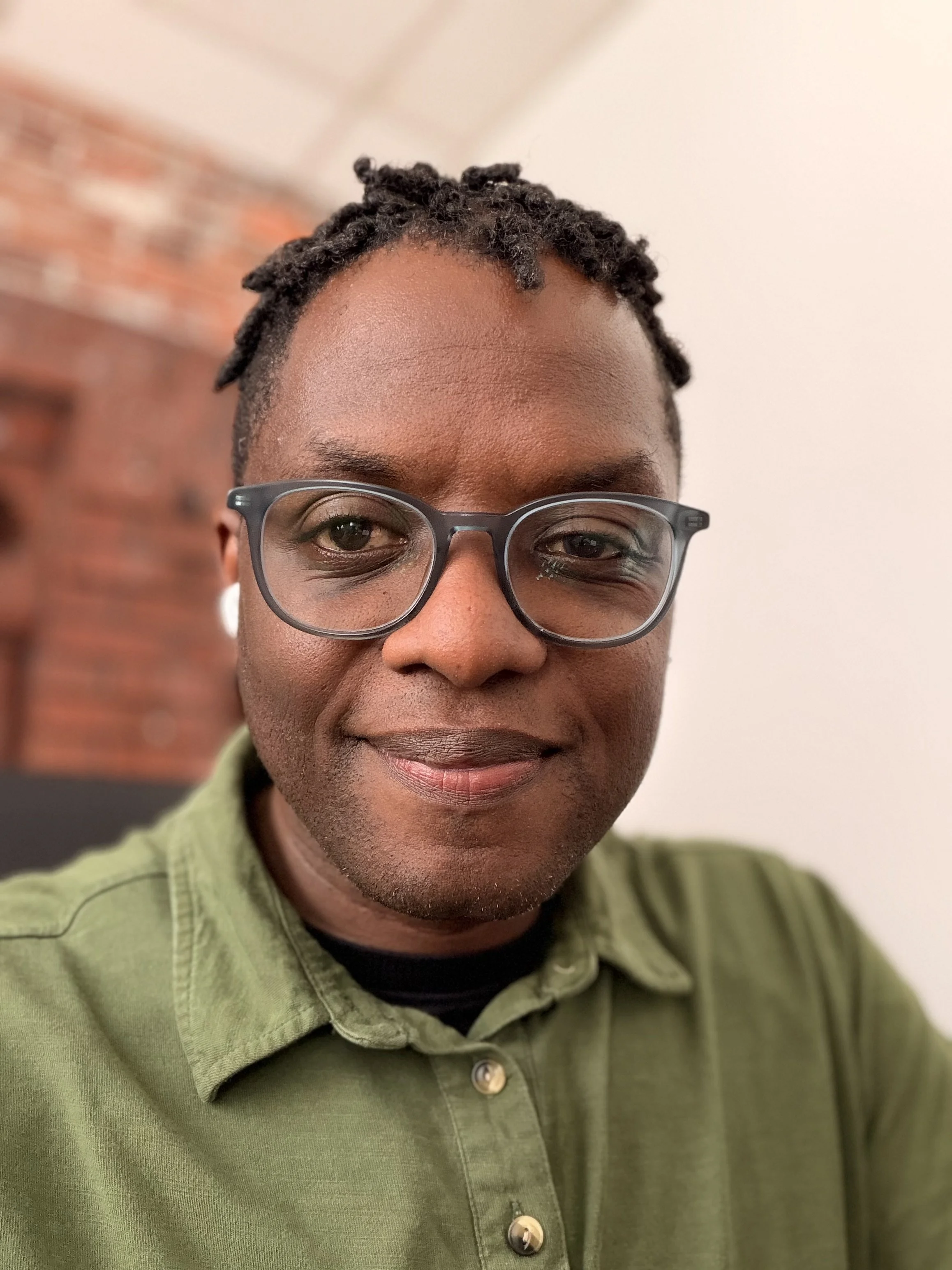 Close-up of a person's face showing the upper half, wearing glasses, with short textured black hair, and dressed in an olive green button-up shirt, against a blurred indoor background.
