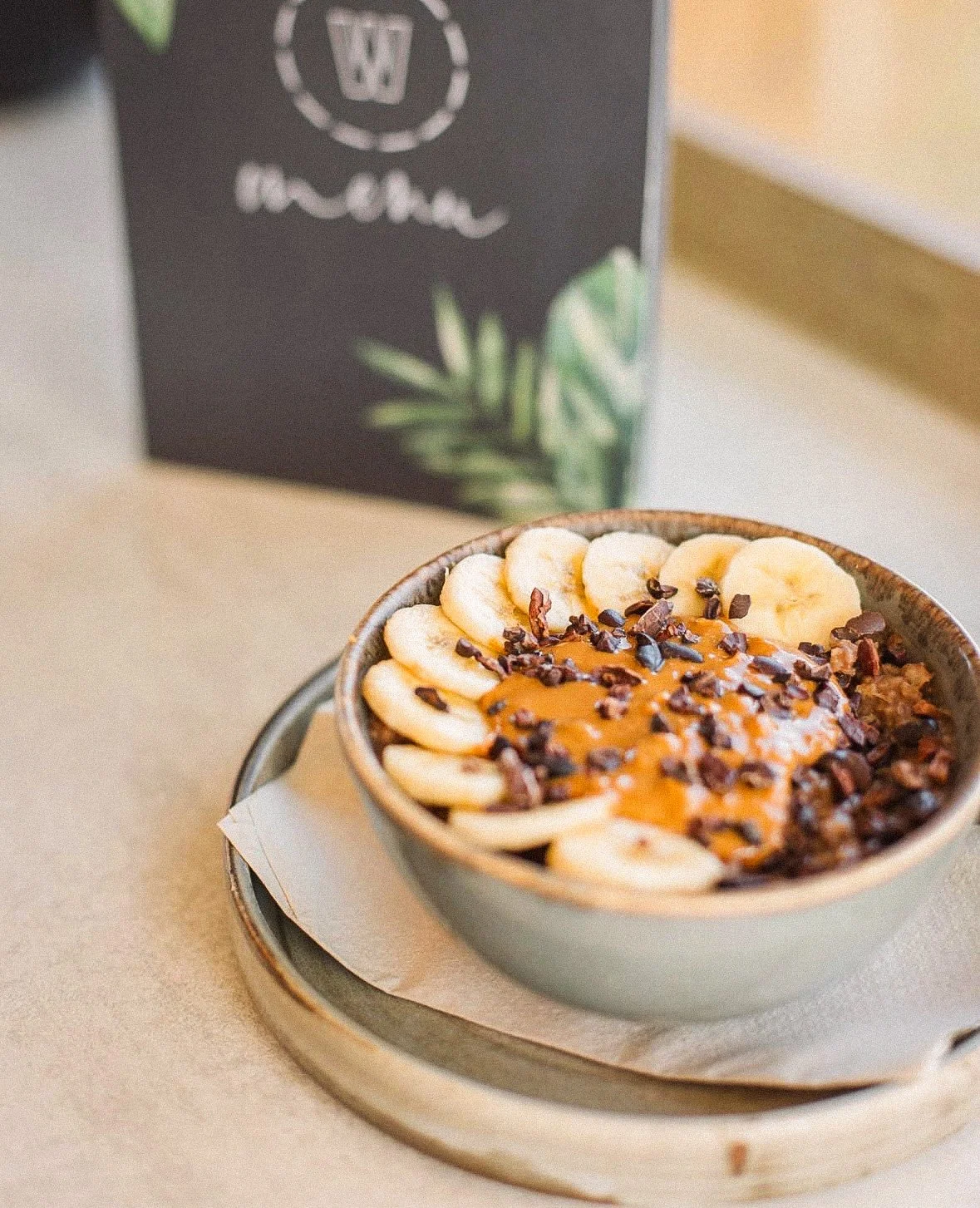 Bowl of oatmeal topped with sliced bananas, chocolate chips, and caramel drizzle on a plate inside a cafe or restaurant setting.
