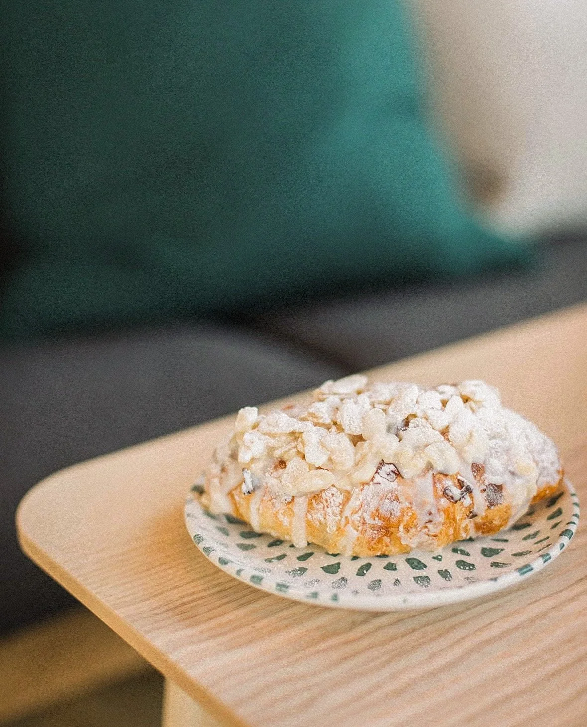 A donut with white icing and chopped nuts on top, placed on a small paper plate with green dot pattern, on a wooden table.