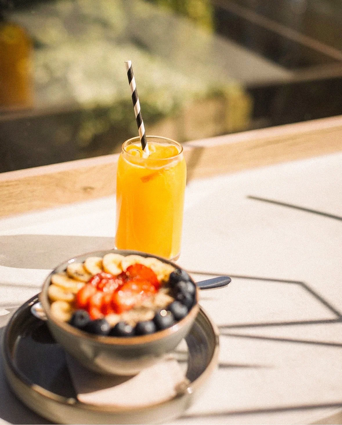 A glass of orange juice with a black and white striped straw and a bowl of fruit topped with strawberries, bananas, and blueberries on a white outdoor table.