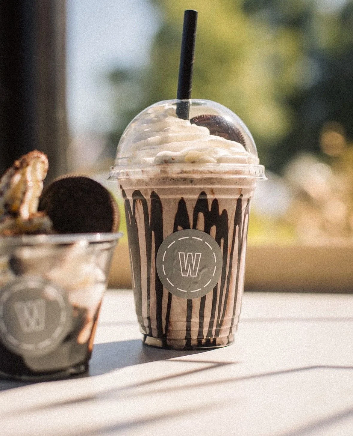 Chocolate milkshake with whipped cream in a clear plastic cup with a black straw and a logo, placed on a white surface with blurred outdoor background.