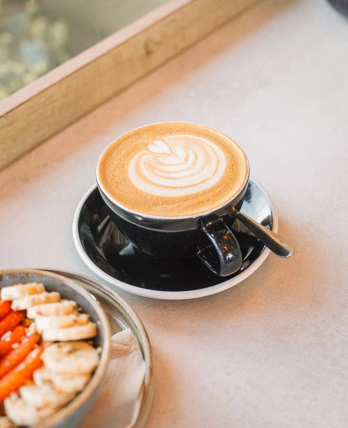 A cup of coffee with latte art on top, placed on a black saucer with a silver spoon resting on it, on a light-colored surface near a window.