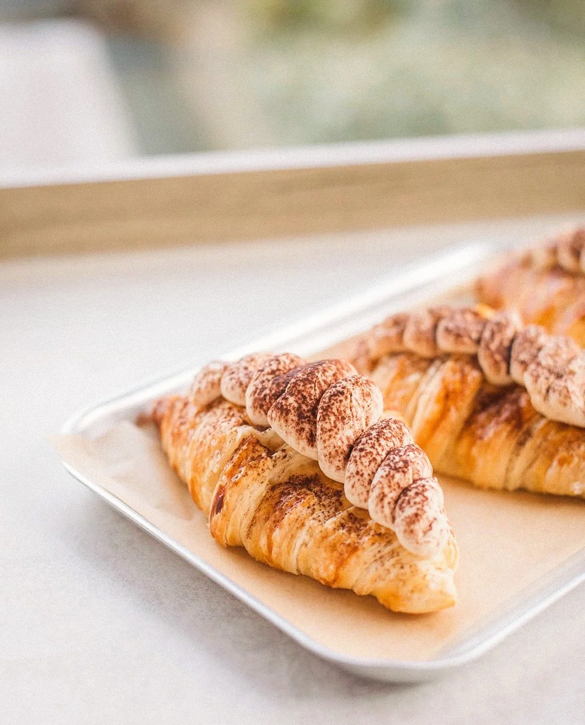 Croissant topped with whipped cream and cocoa powder on a white rectangular plate.