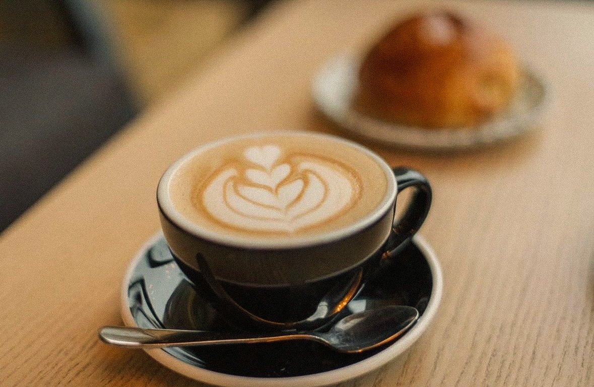 A black cup of latte with latte art on top, placed on a matching saucer with a small spoon, on a light wooden table. In the background, there is a plate with a pastry.
