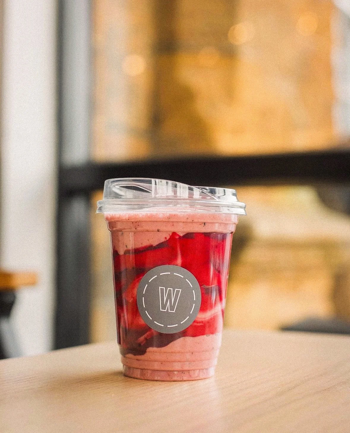 Pink and red layered smoothie in a clear plastic cup with a gray circular logo and a transparent lid, placed on a light wood table.